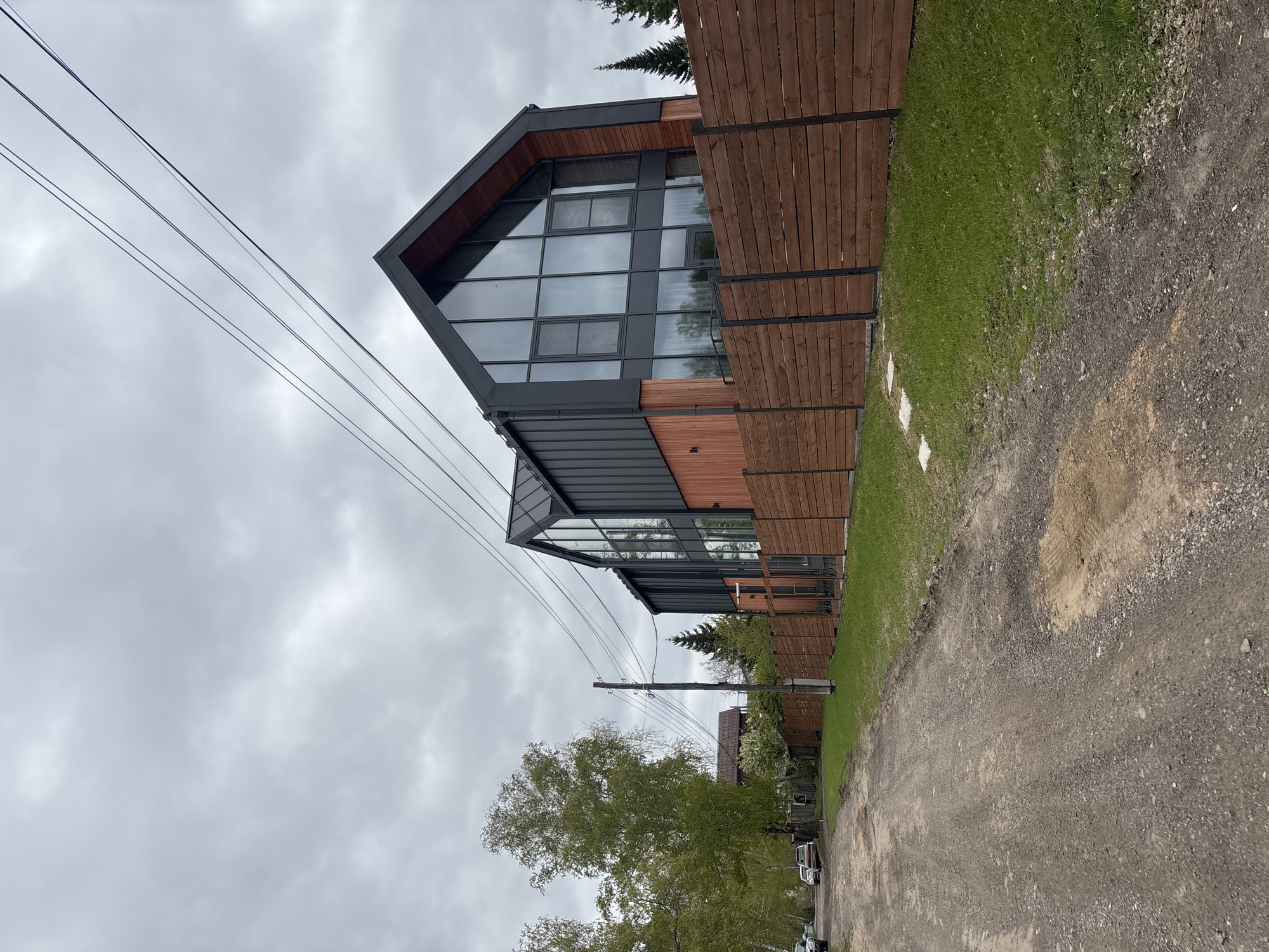 The image depicts a modern residential building in what appears to be a suburban or semi-rural area. The building is characterized by its angular, contemporary design, featuring a large glass facade and dark gray trim, contrasted by sections of wood paneling. A tall, slatted wooden fence runs along the property line adjacent to a gravel or dirt road. Overhead power lines are visible against a cloudy sky. Lush green grass grows between the fence and the road. The setting suggests a location on the outskirts of a city where newer architectural styles are integrated into a landscape that retains elements of a more natural environment. Given the prompt, this would be a possible scene in the city of Krasnoyarsk, Russia. The image is taken from a low angle, emphasizing the height of the building and the overhead power lines. No people or significant activity are visible.