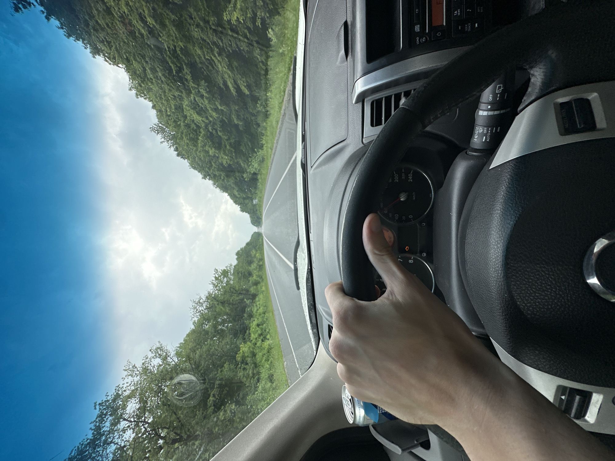 The image shows a first-person perspective from inside a car, with the driver's hands on the steering wheel. The car is on a road, with lush green trees lining both sides of the road. The sky is visible through the windshield, with a mix of blue sky and clouds, suggesting it's daytime. A can with "L-CARNITINE VIT" is visible near the bottom of the frame. The dashboard and steering wheel of the car are also visible. The scene appears to be a road trip through a rural or natural area. The weather seems to be fair, with a blend of sunshine and clouds. The image is oriented sideways.