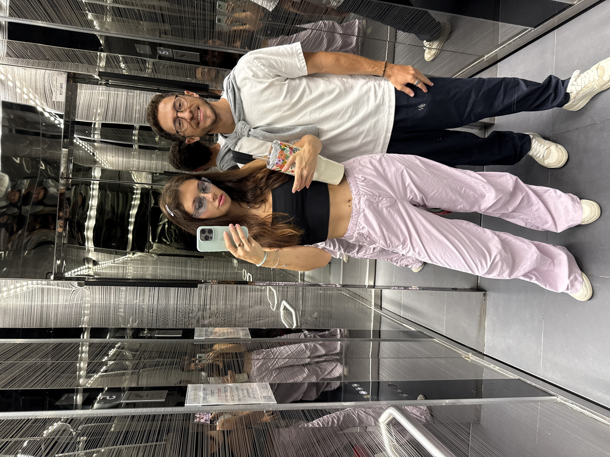 The image shows two individuals, a man and a woman, posing for a selfie in a mirrored elevator. The woman is holding a phone and a cup, while the man is standing behind her. The interior of the elevator has a modern design with multiple mirrors and what appears to be a decorative wall pattern of hanging thin lines. The location is identified as Tel-Aviv, Israel, based on contextual clues not explicitly visible in the image.