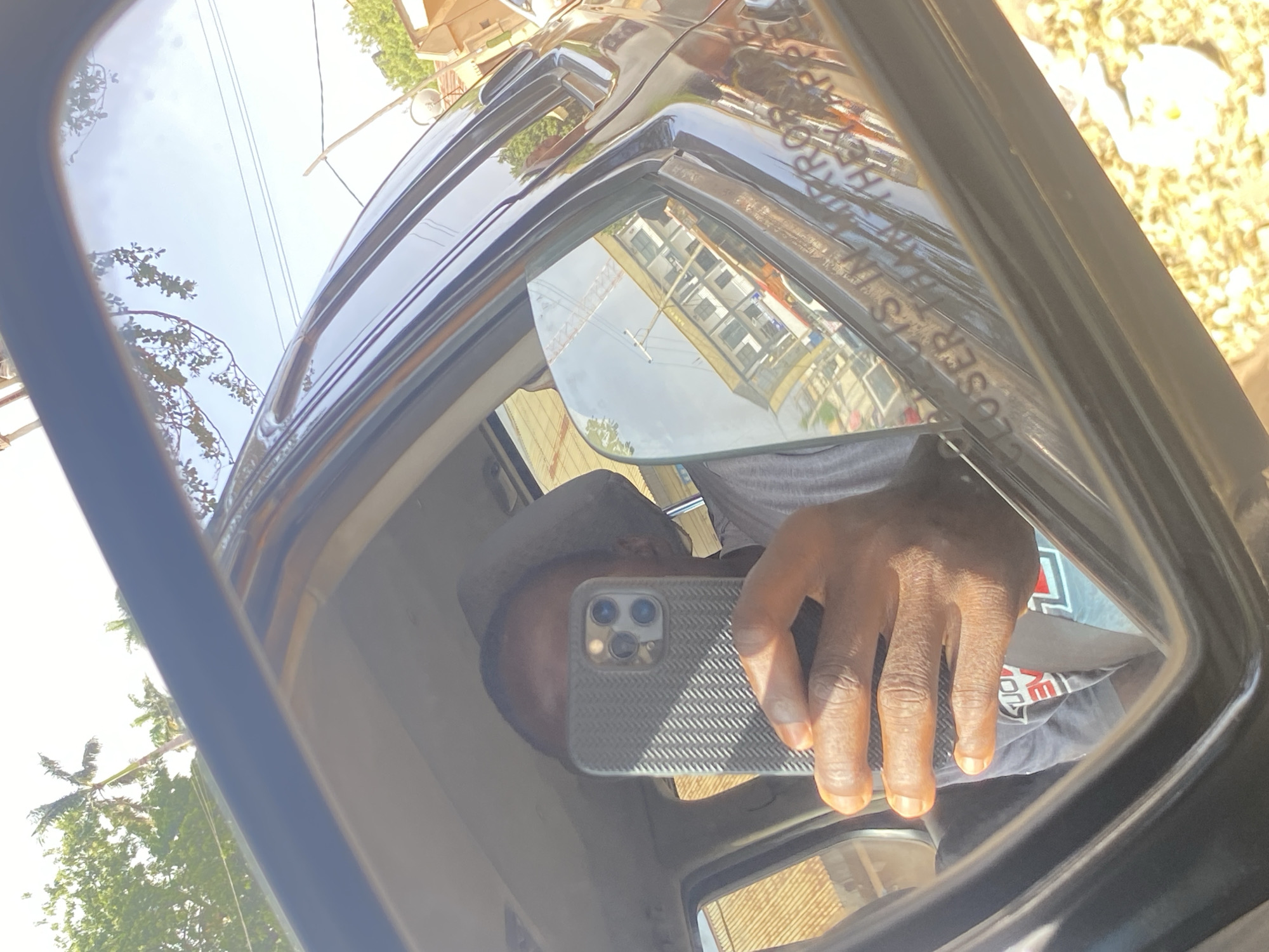 The image displays a bright, daytime selfie captured through the reflection in a car's side mirror. The scene suggests a sunny, outdoor environment.
The primary subject is a person, partially visible as a reflection. Their dark-skinned hand holds a grey smartphone with a textured case and a triple-lens camera system, indicating they are actively taking the photo. Parts of their head, adorned with a grey cap or hood, and a grey t-shirt with red and white graphics are also reflected.
The side mirror and the glossy dark body of the car reflect the surroundings. Above, a clear, bright blue sky with some light haze signifies a sunny day. Green trees and foliage are visible, including what appears to be palm trees. A multi-story building with a light-colored facade and numerous windows is prominently reflected, along with a hint of a construction crane in the distance. Overhead utility lines and a satellite dish are also discernible against the sky. The ground visible in some reflections appears light and dry, possibly unpaved.
A key detail is the mirrored text "OBJECTS IN MIRROR ARE CLOSER THAN THEY APPEAR" visible in the lower right section of the mirror, characteristic of passenger-side vehicle mirrors. The overall bright, warm atmosphere, vegetation, and architecture are consistent with the provided location context of Barden Yana, Nigeria.