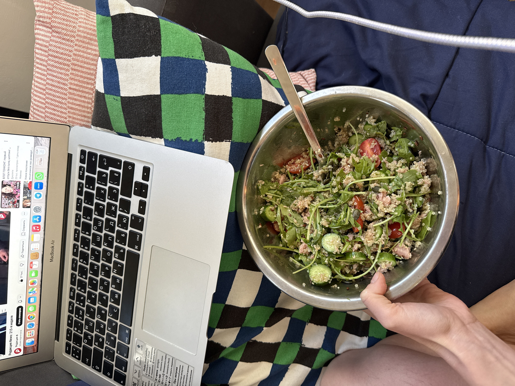 The scene appears to be indoors, likely in a person's home. A person is sitting on a dark blue cushioned surface, possibly a couch or bed, with a laptop and a bowl of salad. The laptop, a silver Macbook Air, is positioned to the left, and the screen displays Russian text and app icons. Next to the laptop is a pillow with a green, white, blue and black checkered pattern. The salad is in a large, silver metal bowl, containing mixed greens, cucumbers, tomatoes, and possibly quinoa or another grain. A silver spoon is stuck into the salad. The person is holding the bowl with their left hand. A white cable is visible in the upper right corner. The overall setting suggests a casual, relaxed environment, possibly during a meal break while working or browsing on the laptop. There is no indication that the location is Tel-Aviv, Israel.