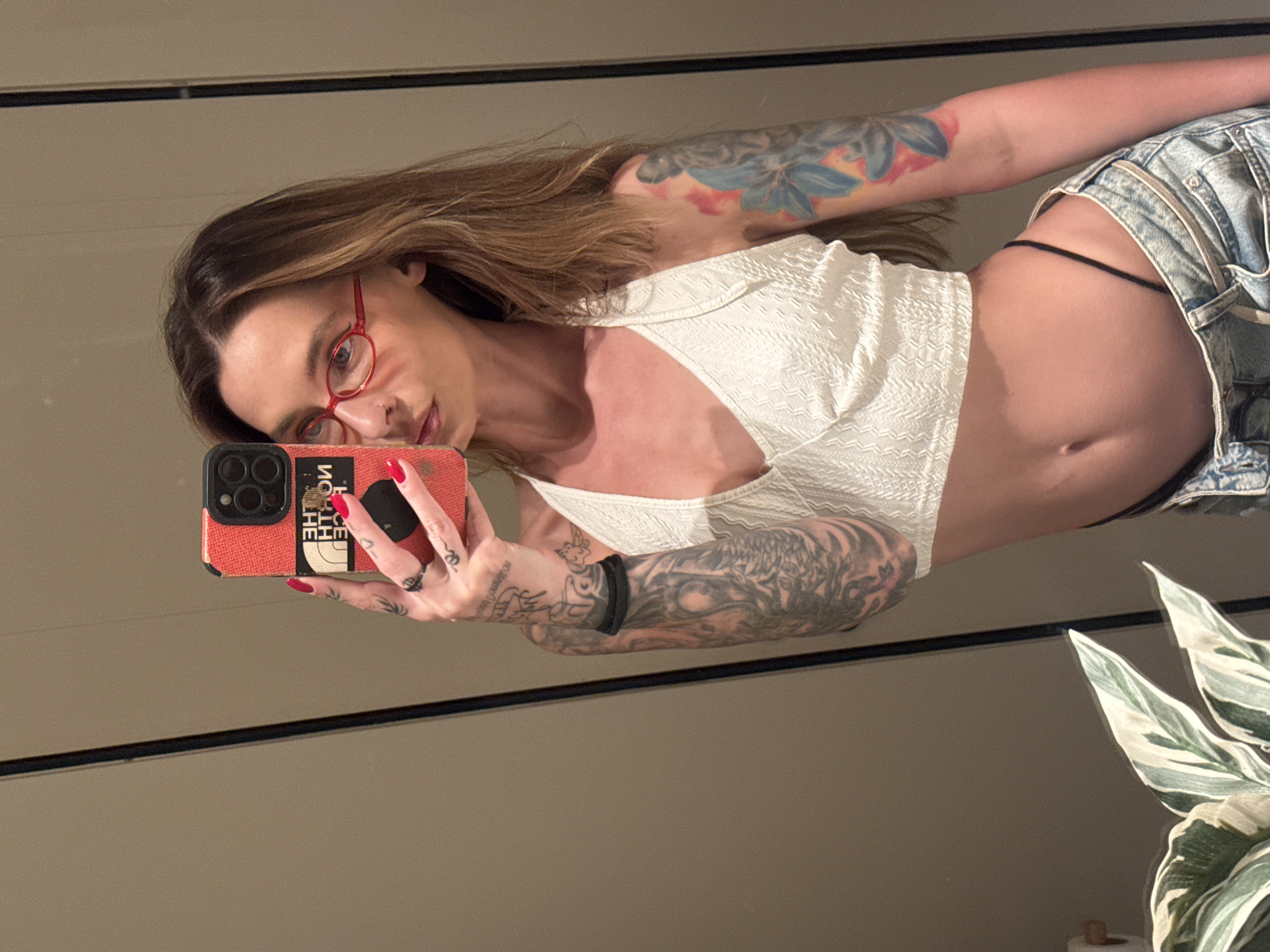 A woman takes a selfie in a mirrored bathroom. She wears red glasses, a white, textured top, and light-wash denim shorts with a visible thin black thong. Tattoos cover both arms. She holds a red and black phone case with the words "THE NORTH FACE." The background includes a wall, a dark horizontal line indicating the mirror's edge, and a houseplant in the corner. The location and city are not discernible from the image.