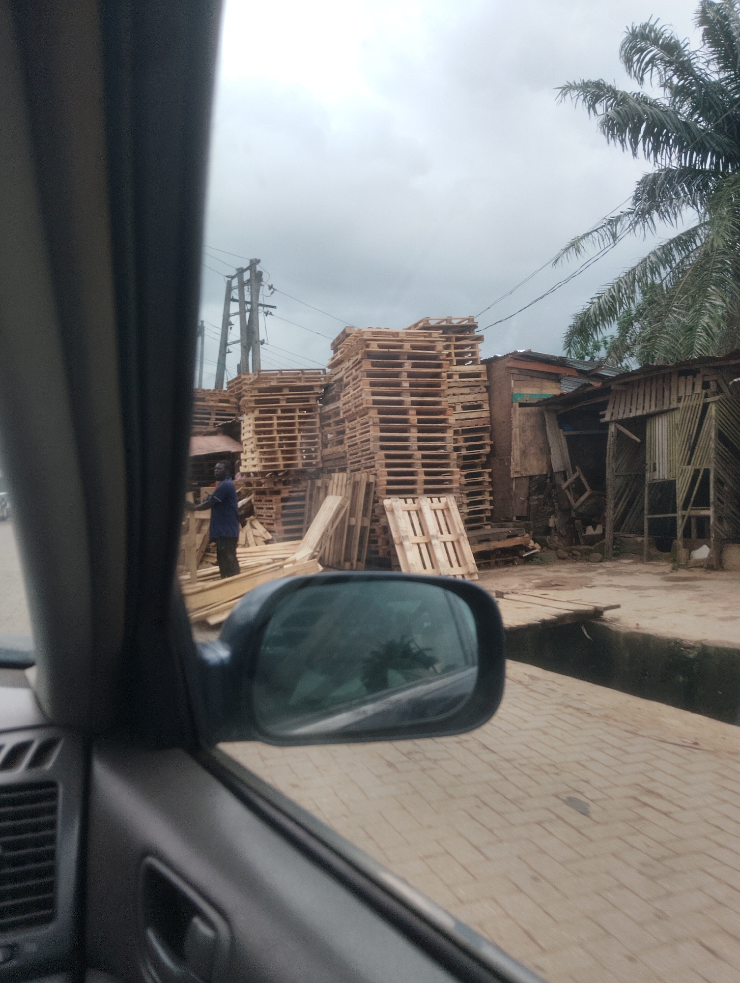 The image shows an outdoor scene in Iju, Nigeria, with a focus on stacked wooden pallets. In the foreground, the interior of a car is visible, including the side mirror, dashboard, and door panel. Through the side window, a man in a blue shirt and dark pants is standing among stacks of wooden pallets. He appears to be interacting with the pallets. Behind the man, large stacks of wooden pallets are piled high, suggesting a storage or distribution area for these items. A palm tree is visible on the right side of the image. Power lines and utility poles are present in the background, indicating an urban or semi-urban environment. The ground appears to be paved with interlocking bricks. The sky is overcast, suggesting a cloudy day.