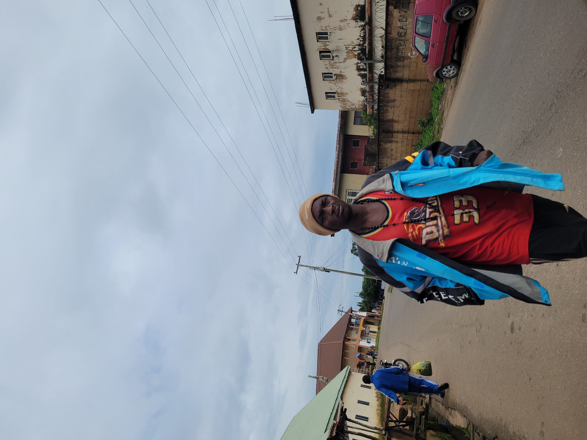 A medium shot shows a young Black man walking towards the camera on a paved street in Jos, Nigeria. He is wearing a beige beanie, a red tank top with yellow and black detailing and the number "33" on it, and a light blue and grey jacket. He has a beaded necklace. He is in the foreground, with his body angled slightly to his right. In the background, a man in a blue jacket is walking away from the camera, carrying a green bag. Several buildings line the street, including a light-colored building with a green roof on the left and a beige building with peeling paint and barred windows on the right. A red car is parked on the side of the road to the right of the frame. Overhead, power lines are visible against a cloudy sky.