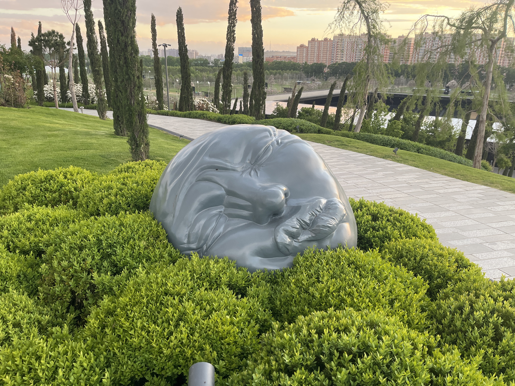 The image depicts a gray sculpture of a sleeping person nestled in green bushes, located in a park. A paved pathway runs adjacent to the sculpture. In the background, there is a row of tall, thin trees, followed by a body of water with a bridge visible in the distance. Buildings are also seen in the far background. The park is in Krasnodar, Russia.
