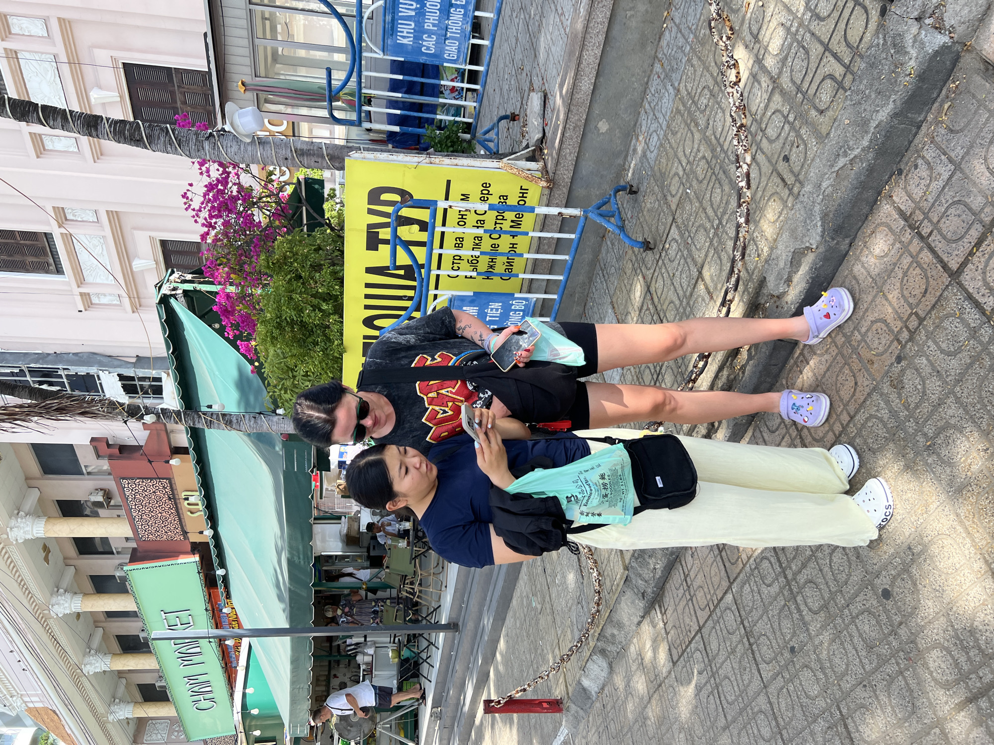 Two women are standing on a sidewalk in Nha Trang, Vietnam, looking at a smartphone. The woman on the left is wearing a navy blue t-shirt and light yellow wide-leg pants. She is holding a light blue plastic shopping bag in her left hand. The woman on the right is wearing a black t-shirt with a graphic print and black shorts. Both women are wearing casual shoes. Behind them, there is a building with a green awning and a sign that reads "CHAM MARKET." There are also palm trees and a yellow banner with text that is partially obscured. The sidewalk is paved with patterned tiles, and there is a metal chain link fence in the background. The overall scene suggests a commercial area in a city.