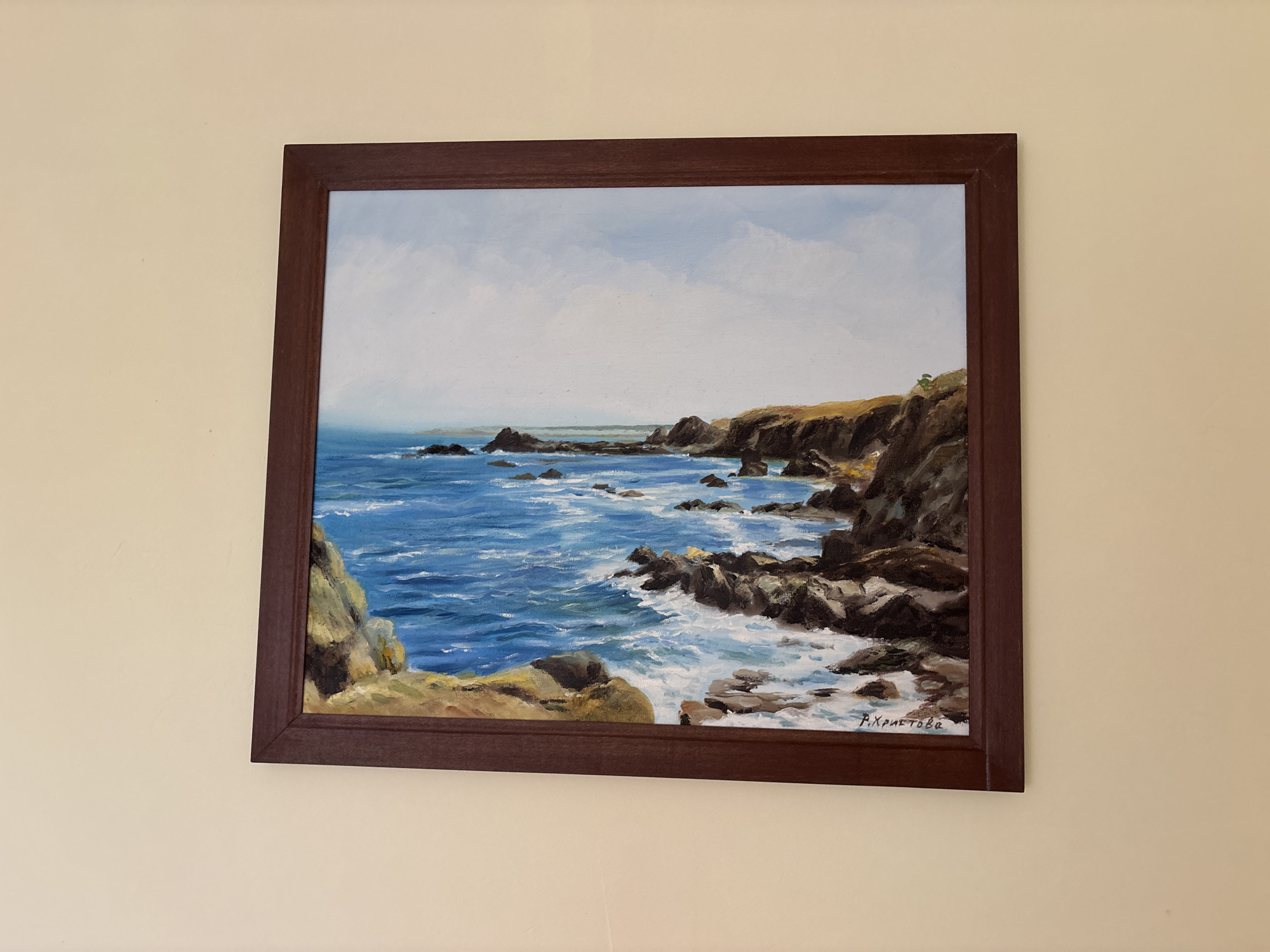 A framed oil painting hangs on a beige wall, depicting a coastal landscape with rocky shores and the sea. The painting is oriented horizontally, showcasing a dynamic scene of waves crashing against dark, rugged rocks and cliffs. The water is a vibrant blue, with white foam indicating the force of the waves. The cliffs are rendered in shades of brown and green, with sparse vegetation clinging to their surfaces. The sky above is a soft blue with scattered white clouds. In the lower right corner of the painting, a signature in cursive script reads "P. Христова," which is likely the artist's name. The painting itself is enclosed in a dark brown frame. The scene is characteristic of the coastline in Rezovo, Bulgaria, a town known for its dramatic sea cliffs and natural beauty. There are no people or animals depicted in the painting. The image is a close-up shot of the artwork on the wall, with the wall and frame being the only visible elements outside of the painting itself.