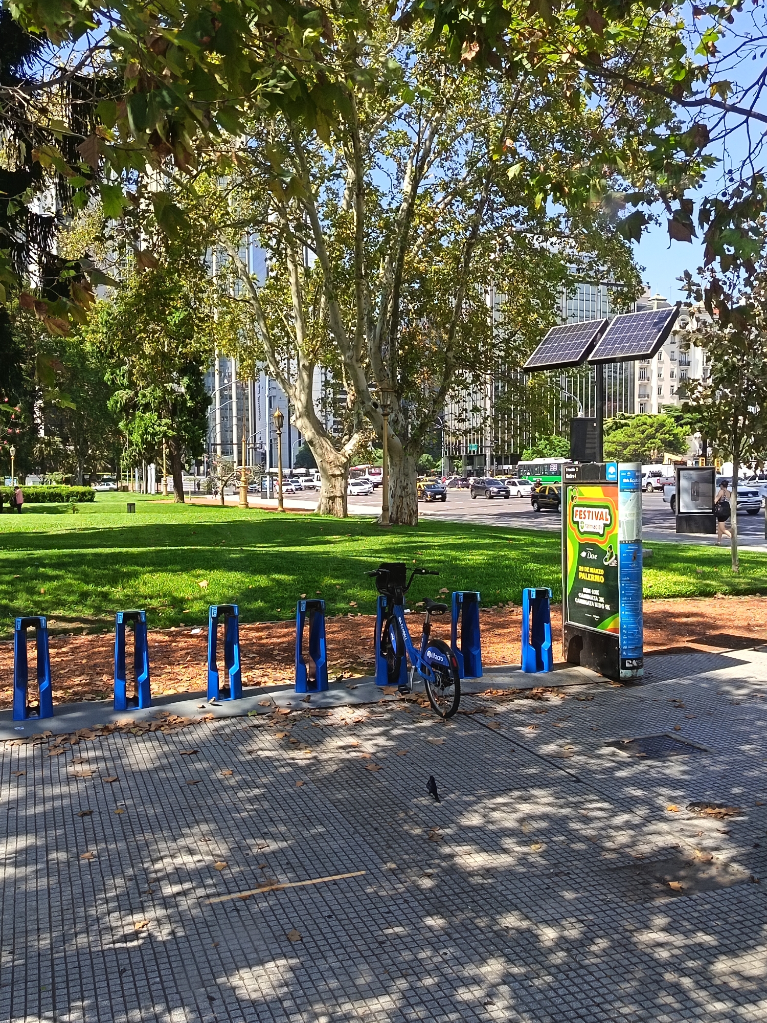 The image shows a bicycle sharing station in a park in Buenos Aires, Argentina. A blue bicycle is parked at one of the docking stations. Behind the station is a green lawn with large trees. Across the street, there is traffic and buildings. A person is walking on the sidewalk in the background. The weather appears to be sunny. The advertisement on the station is for the Festival Farmacity on March 20th in Palermo. There are solar panels atop the kiosk.