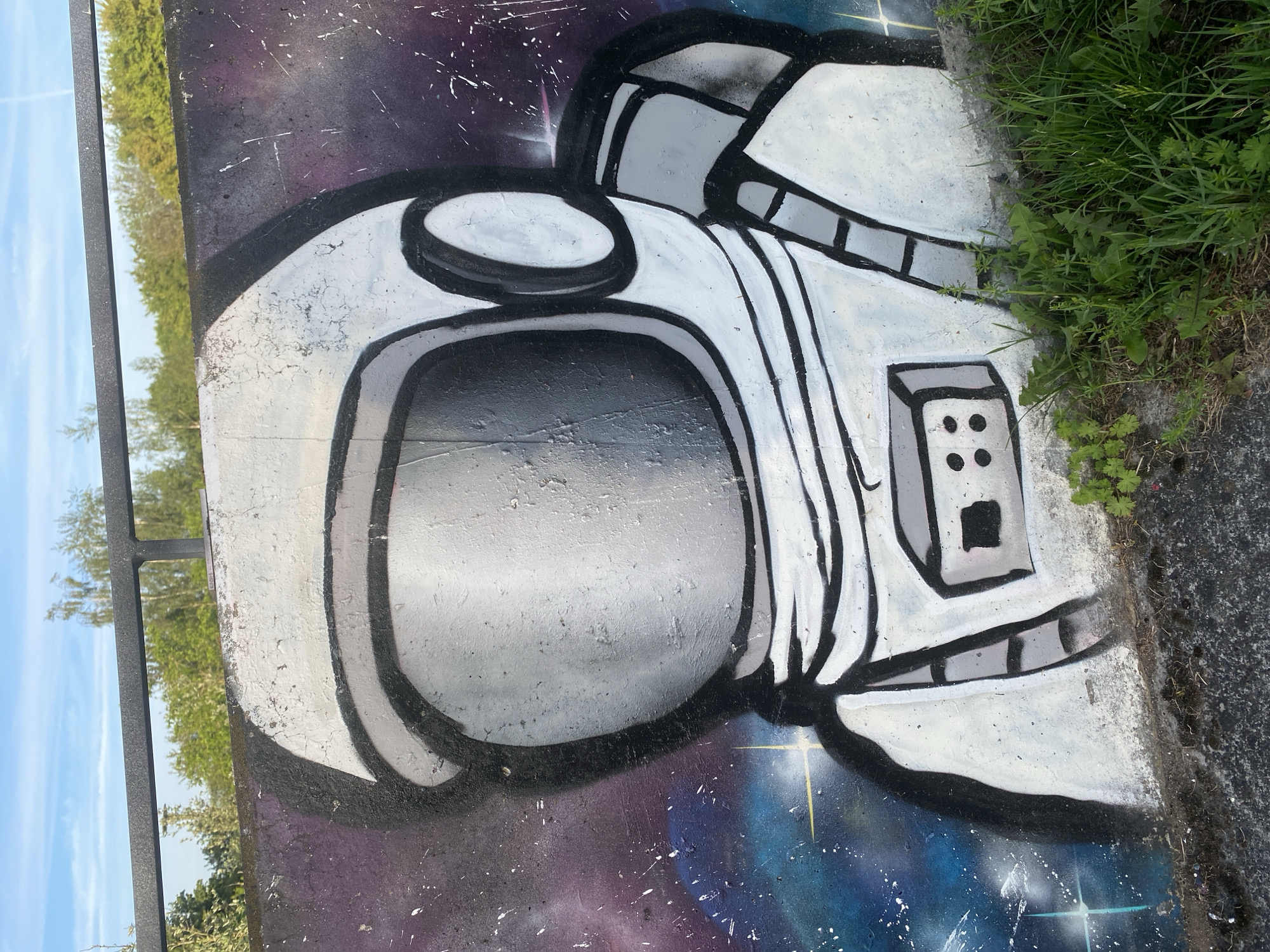 A graffiti mural depicting an astronaut's helmet is painted on a concrete wall. The helmet is predominantly white with black outlines and grey shading to create a three-dimensional effect. The visor is a reflective grey, and the backdrop is a spray-painted space scene with shades of purple and blue and scattered white specks representing stars. Patches of green grass and weeds are growing along the edge of the concrete wall. A metal railing and a glimpse of trees and sky are visible to the left of the mural. The location is Landau in der Pfalz, Germany.
