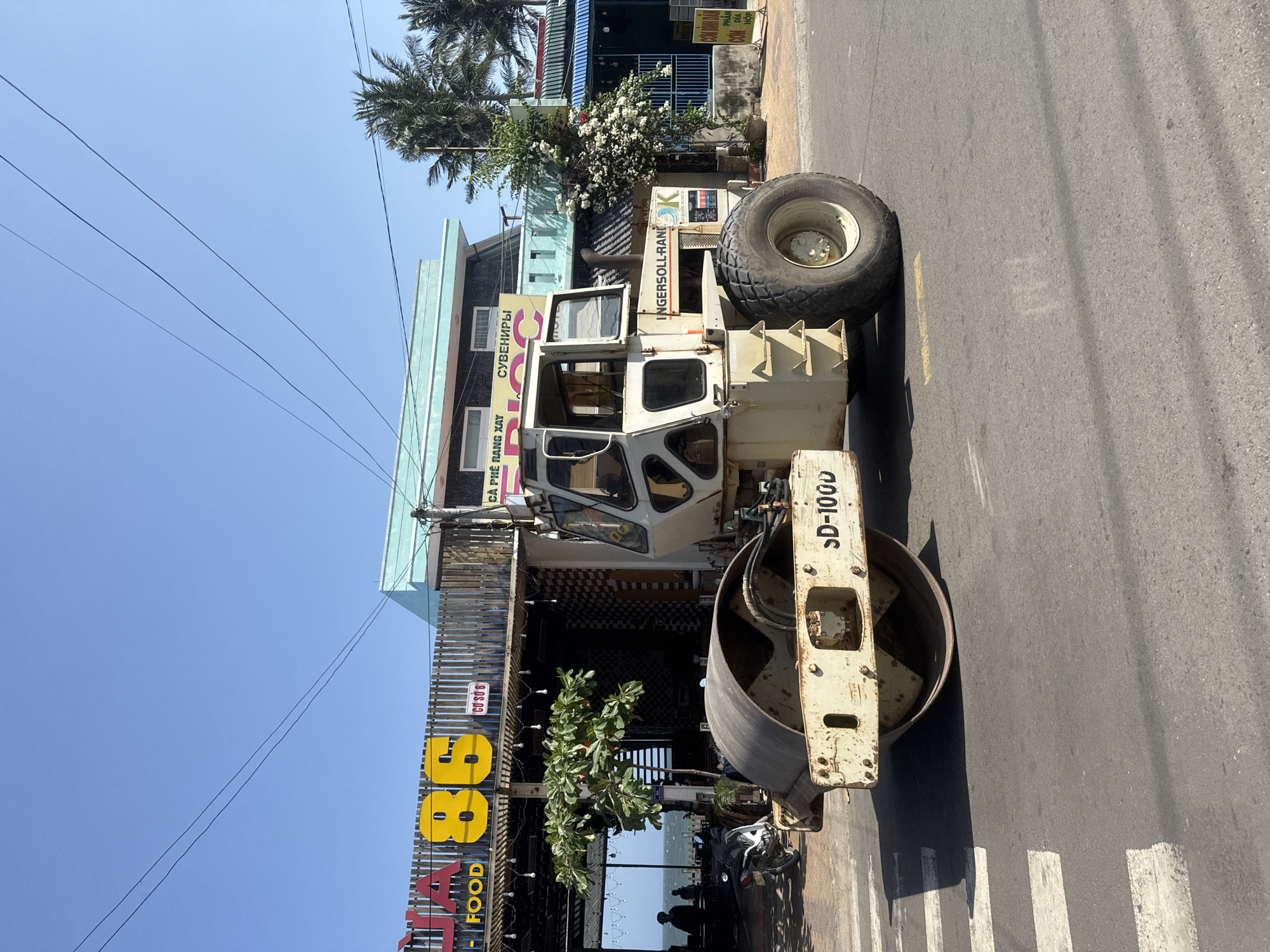 In Mũi Né, Vietnam, a large, yellow and white Ingersoll-Rand SD-1000 road roller is parked on an asphalt road in front of a beachfront establishment with a prominent yellow sign displaying "86 FOOD". The road roller is the primary subject, its heavy machinery dominating the foreground. Behind it, a building with a light turquoise upper floor and darker textured lower facade is visible. A wooden fence separates the building from the street. Palm trees are scattered in the background, suggesting a tropical coastal environment. A motorcycle is parked to the left of the road roller. The sky is clear and bright blue. A pedestrian crossing with white stripes is visible on the road in front of the roller. The scene appears to be a street in a commercial area near the beach.