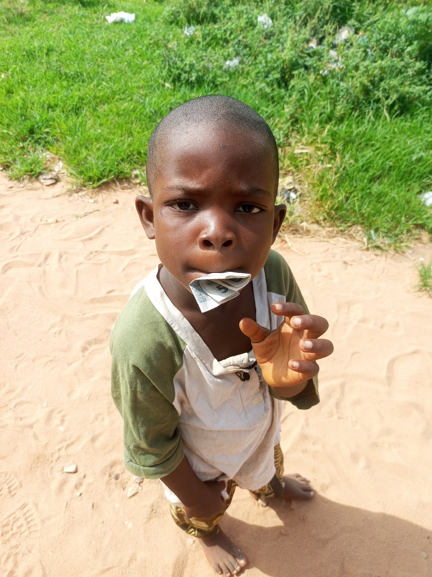 A young Black child, appearing to be around 5-6 years old, is pictured from a high-angle shot looking upwards. The child has dark skin and a shaved head. They are wearing a light-colored shirt with a green collar and sleeves, and a matching bib-like garment over it. The child is holding a folded banknote in their mouth and their right hand is raised, with fingers slightly curled. The child is standing on a sandy surface, with footprints visible. In the background, there is green grass and some scattered white debris. The scene appears to be outdoors in an unpaved area. The lighting suggests daytime. There is no information in the image to determine the location as Tempure, Nigeria.