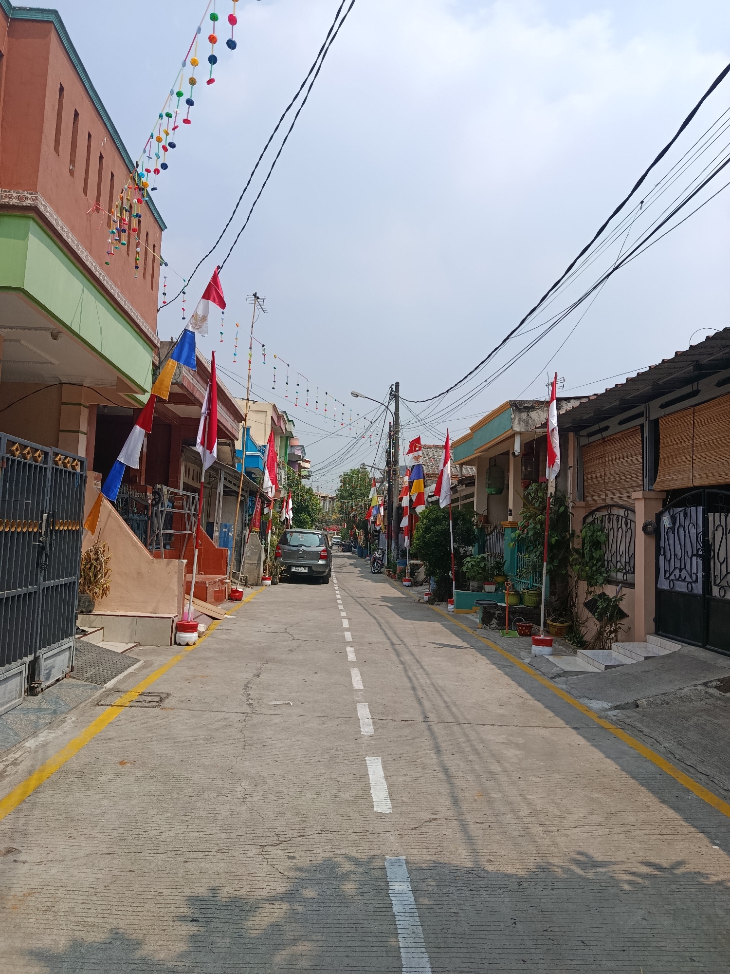The image captures a street scene in Wanasari, Indonesia, decorated with numerous Indonesian flags and colorful festive banners. The flags, predominantly red and white, are affixed to poles along the sides of the street and are also strung across the road. The banners, consisting of small, circular, multi-colored elements, hang overhead.
The street itself is paved and features a dashed white line marking the center. Yellow lines delineate the edges of the road. On either side of the street are residential buildings with distinct architectural features, including colorful facades, window grills, and various plant life. Some of the buildings have gates made of metal bars.
A dark-colored hatchback car is parked on the right side of the street. Power lines and utility poles are visible overhead, crisscrossing the sky. The sky is bright and appears to be partly cloudy, suggesting daytime. The overall impression is that of a residential area prepared for a celebration or national holiday.