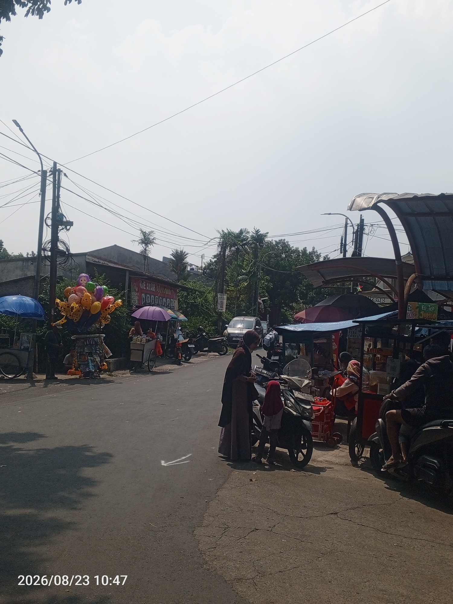 The image shows a street scene in Depok, Indonesia, on August 23, 2026, at 10:47 AM. The sky is overcast. On the left side of the road, there are vendors selling balloons and other items. One vendor has a cart full of colorful balloons, including some with animal prints. Another vendor has a blue umbrella. Across the street, there are more vendors with carts and umbrellas, selling food and drinks. Several motorcycles are parked along the roadside, and some people are interacting with the vendors. A silver car is visible in the background. The road is paved, and there are trees and buildings lining the street. The overall impression is of a bustling street market or roadside commerce.