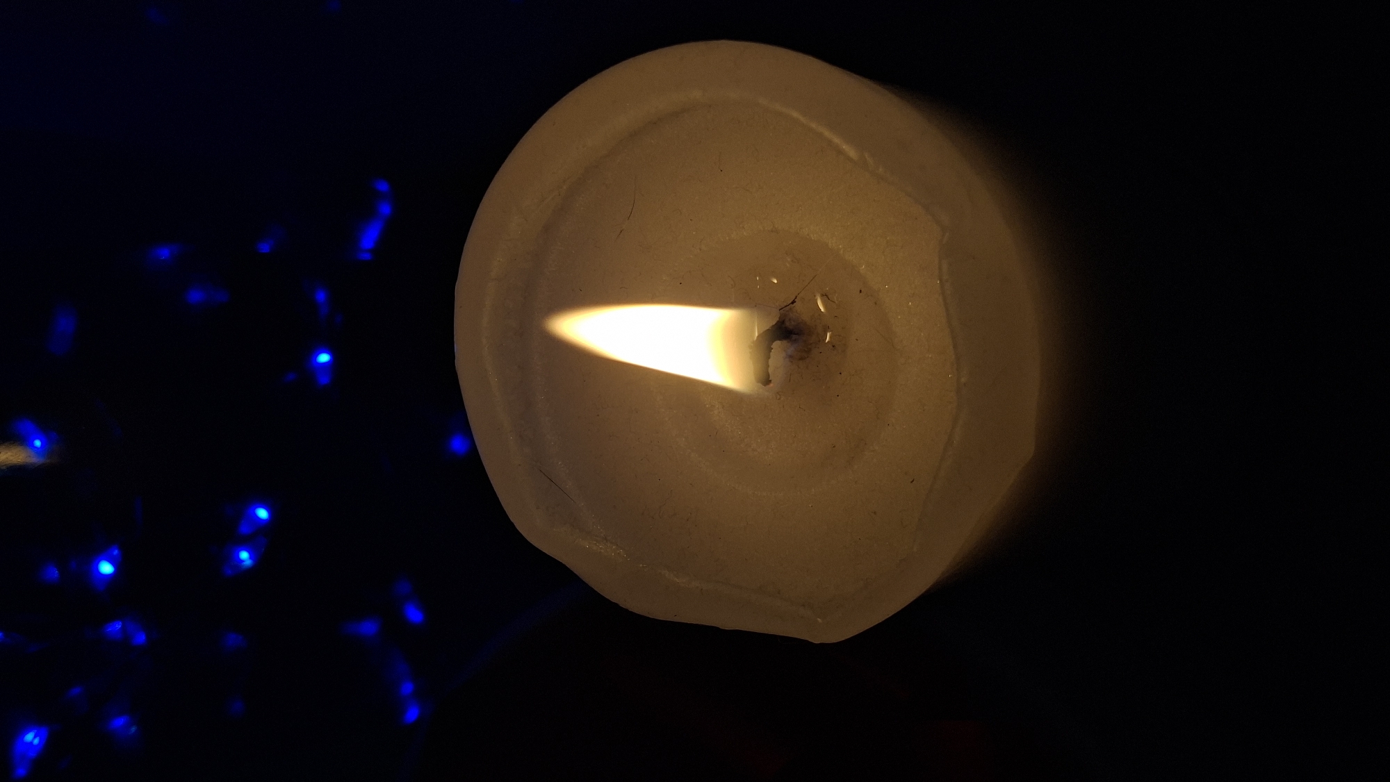 A close-up, low-angle shot captures a lit candle with a yellow flame against a dark background. The candle is a pale, off-white color and appears to be made of wax, with an irregular, textured surface. The flame is bright and flickers upwards, casting a warm glow on the wax. In the upper portion of the image, blurred blue lights are visible, creating a bokeh effect. The lighting suggests a dim, possibly indoor environment. The scene is located in Забалоцкі сельскі Савет, Belarus. There are no people or animals visible in the image.