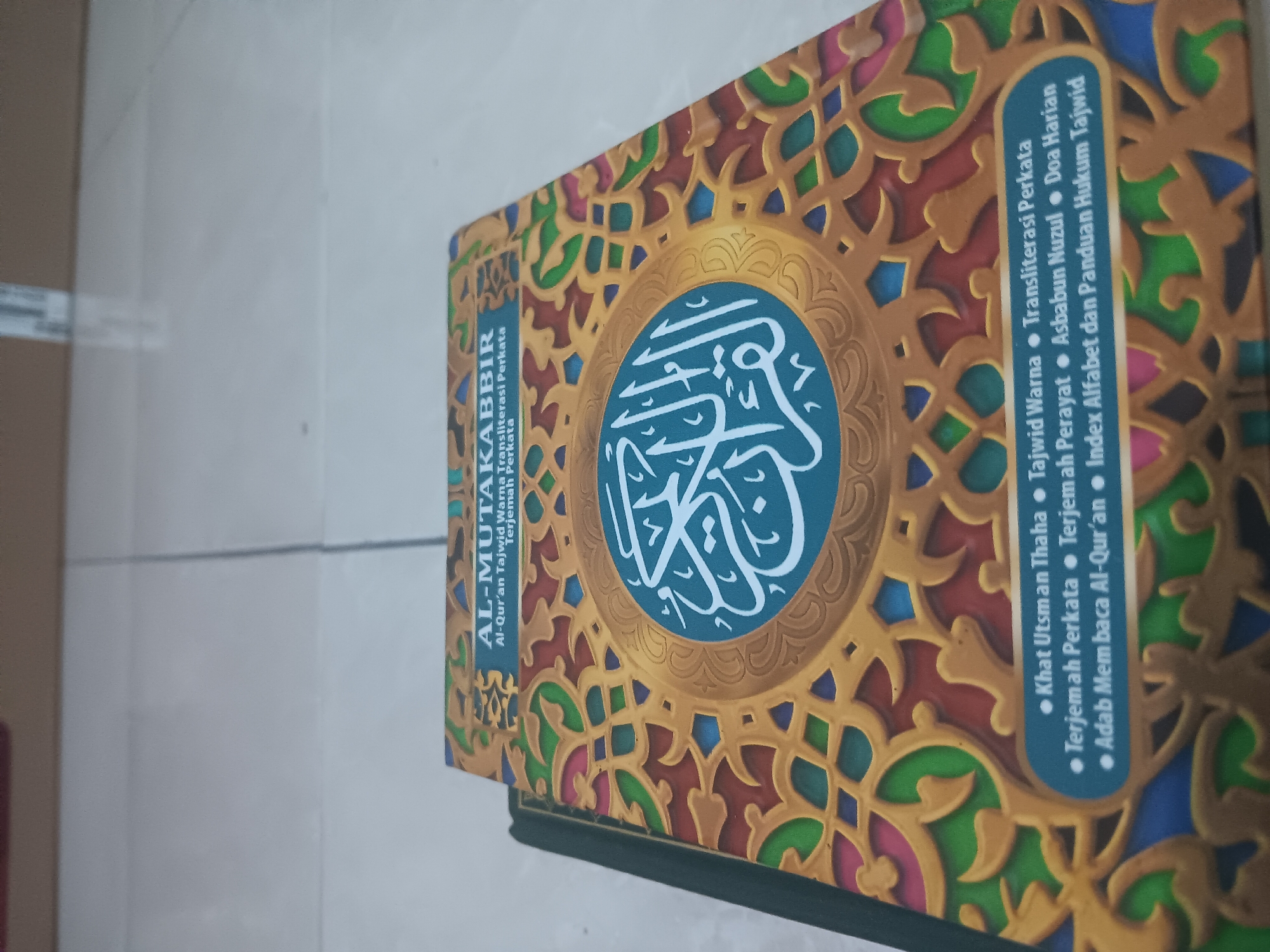 The image shows a close-up view of an open book with ornate decorations on its cover, placed on a light-colored tiled floor. The book's cover features intricate geometric patterns in gold, green, red, and blue. In the center of the cover is a circular emblem with white Arabic calligraphy. The title of the book, "AL-MUTA'AKKABIR," is visible at the top of the cover, followed by smaller text in Indonesian: "Al-Qur'an Tajwid Warna Transliterasi Perkatar Terjemah Perkatar". Below this, a list of features is presented in bullet points with dots as separators: "Khat Utsman Thaha," "Tajwid Warna," "Transliterasi Perkatar," "Terjemah Perkatar," "Terjemah Perayat," "Asbabun Nuzul," "Doa Harian," "Adab Membaca Al-Qur'an," and "Index Alfabet dan Panduan Hukum Tajwid." The book is propped open, revealing its interior. The background consists of a tiled floor with visible grout lines, suggesting an indoor setting. There are no people visible in the image. The lighting appears to be from above, casting a slight shadow from the book onto the floor. The overall scene is static and focused on the book. The image does not contain any information that would allow for the determination of its location in Batursari, Indonesia.