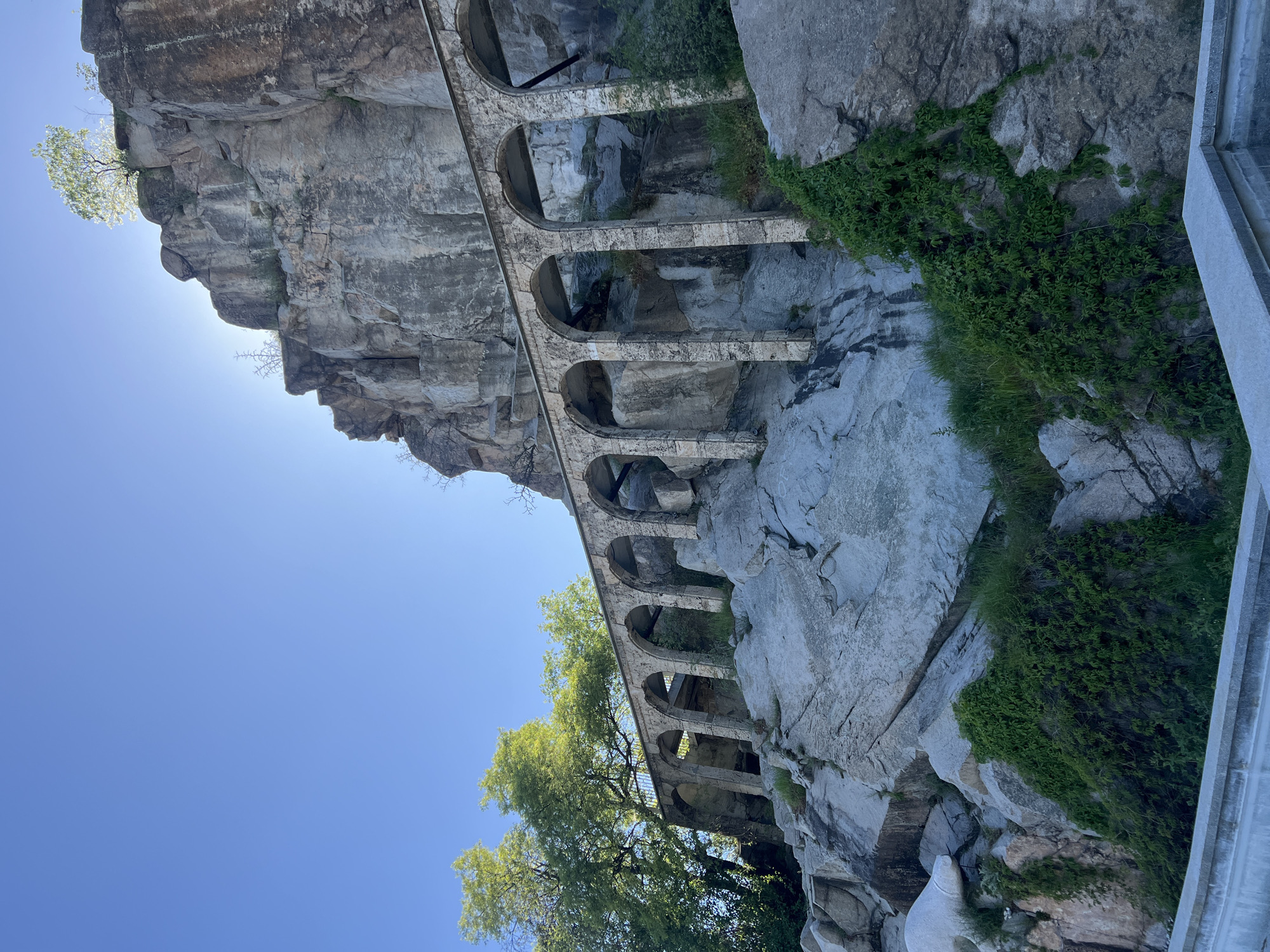 The image depicts the ancient Roman aqueduct, located in Plovdiv, Bulgaria. The aqueduct is constructed of light-colored stone and features a series of arches. It is built against a sheer rock face. Some greenery is visible on the rocks. The sky is clear and blue.