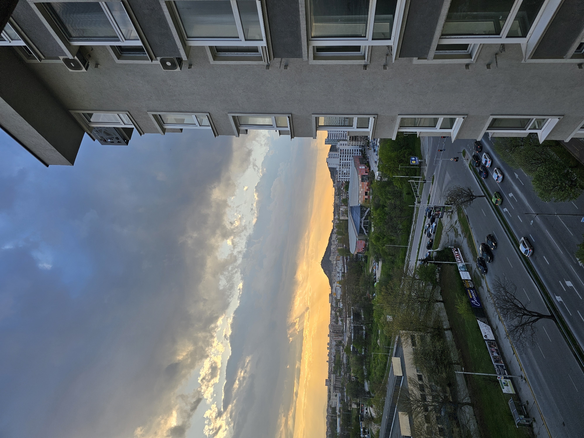 An upward-angled view shows a street scene in Plovdiv, Bulgaria, between two tall buildings. The street, lined with trees, billboards, and parked cars, has a stream of vehicles moving on it. In the distance, a cityscape is visible under a sky that transitions from a cloudy, dark blue to a lighter, sunlit gradient. The architecture of the buildings flanking the view suggests an urban environment.