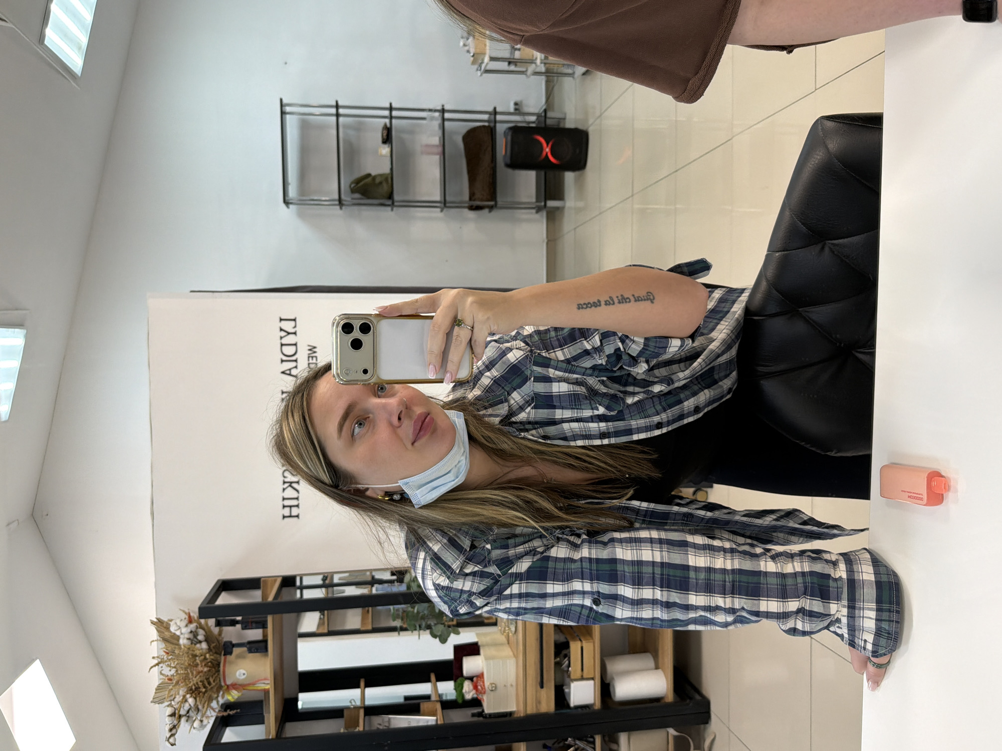 A young woman is captured in a mirror selfie inside what appears to be a modern commercial interior, likely a salon or beauty studio, in Chaikovsky, Russia. The image is oriented vertically, showing the woman's reflection. She has long, light brown hair with blonde highlights and is wearing a blue and white plaid flannel shirt over a dark top. A light blue surgical mask rests below her chin. She holds a silver-colored smartphone with a clear case and a prominent camera array, angled towards the mirror to take the photo. A tattoo is visible on her right forearm, displaying reversed text that reads "lacca ol ih lucca". She wears a ring on her left hand and has manicured nails.
The background, also reflected in the mirror, reveals several features of the establishment. To her right, a black, tufted, high-back chair, typical of a salon, is visible. Behind her, a black metal shelving unit holds various items, including a brown object resembling a boot, a dark green bag, and a black portable speaker with a prominent red 'X' logo. Further to her left, another black shelving unit displays dried floral arrangements, including cotton bolls and grasses, alongside stacked white rolls, possibly towels or paper products. The wall behind her is white and features reversed black text, partially visible as "IHYIAD" and "HIK". On a white surface to her left, a small, light orange rectangular object, resembling a disposable vape pen, shows the reversed text "X000000". The bright, clean environment is illuminated by overhead lighting, casting reflections in the mirror.