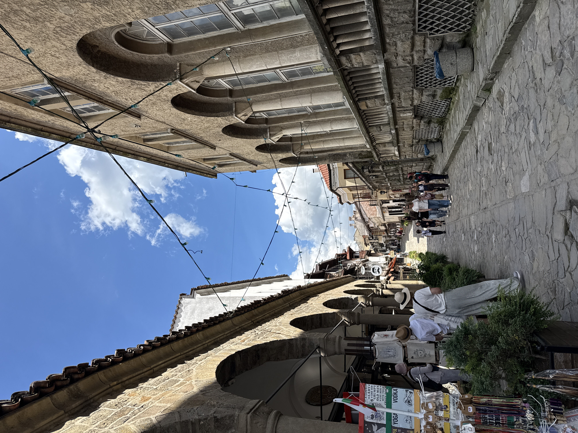 The image captures a street scene in Veliko Tarnovo, Bulgaria. The perspective is from the ground looking up at an angle, showcasing a narrow cobblestone street flanked by historic buildings. Buildings are adorned with arches and windows, reflecting the city's architectural heritage. Overhead, a string of lights is suspended across the street.
In the foreground, a vendor is displaying merchandise, with tote bags, scarves, and souvenirs on display. The vendor stands behind the display with a white brimmed hat. There is also a flag that is red, green, and white. The vendor is positioned near some greenery in the lower right corner of the image.
Along the street, pedestrians can be seen walking in the distance, and the sky is blue with scattered clouds.