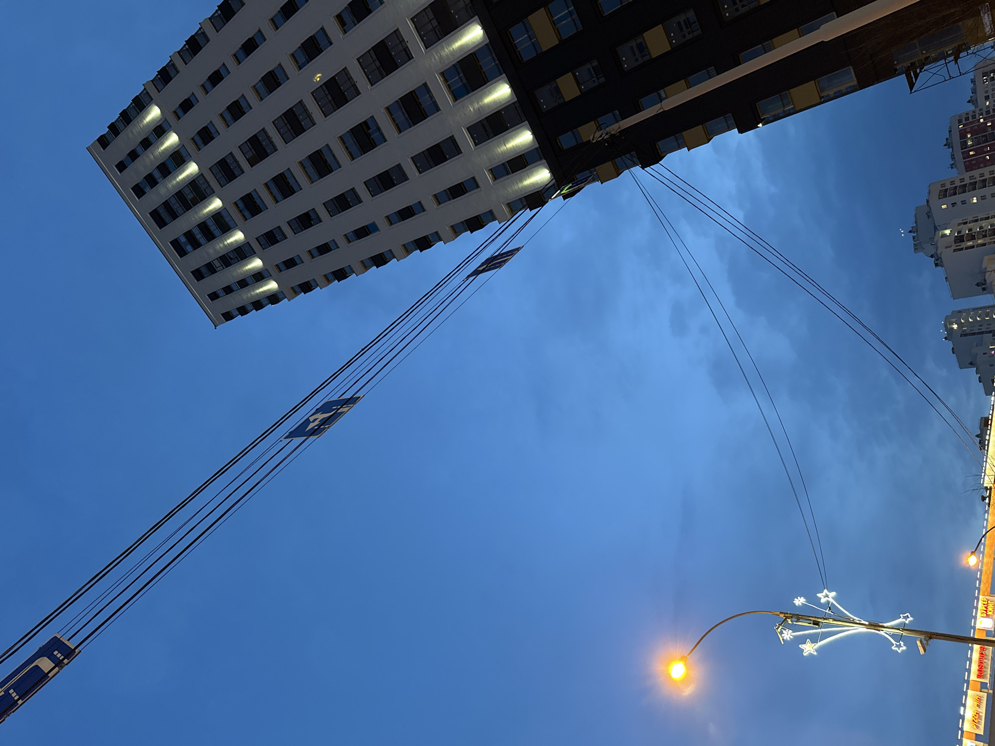 The image shows a low-angle view of a building in Yekaterinburg, Russia at dusk. The building is tall, white, and modern with many windows. A portion of the building is black. Electrical lines stretch across the frame, connecting to the building and to a street lamp in the foreground. The street lamp is illuminated and has a decorative starburst design. Other buildings can be seen in the distance to the right. The sky is a dark blue color.
