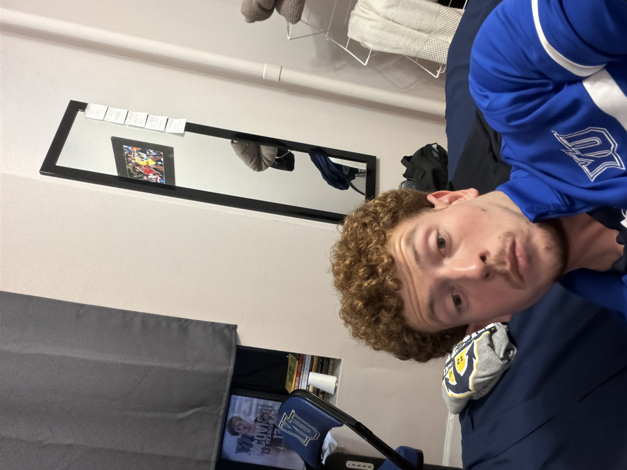 A person with curly brown hair is positioned in the lower right of the frame, wearing a blue jacket with white accents. The person's face is visible. Behind the person, a dresser with a gray covering is partially visible, along with a picture frame bearing the logo "YU" and an image of a basketball player in the lower left. In the background, a black-framed mirror hangs on a light-colored wall. In the mirror's reflection, a picture frame and hanging clothes are visible. Above the mirror, a white pipe runs horizontally across the wall. The photograph was most likely taken in a dorm room and due to the “YU” logo, the location is most likely New York City, United States of America.