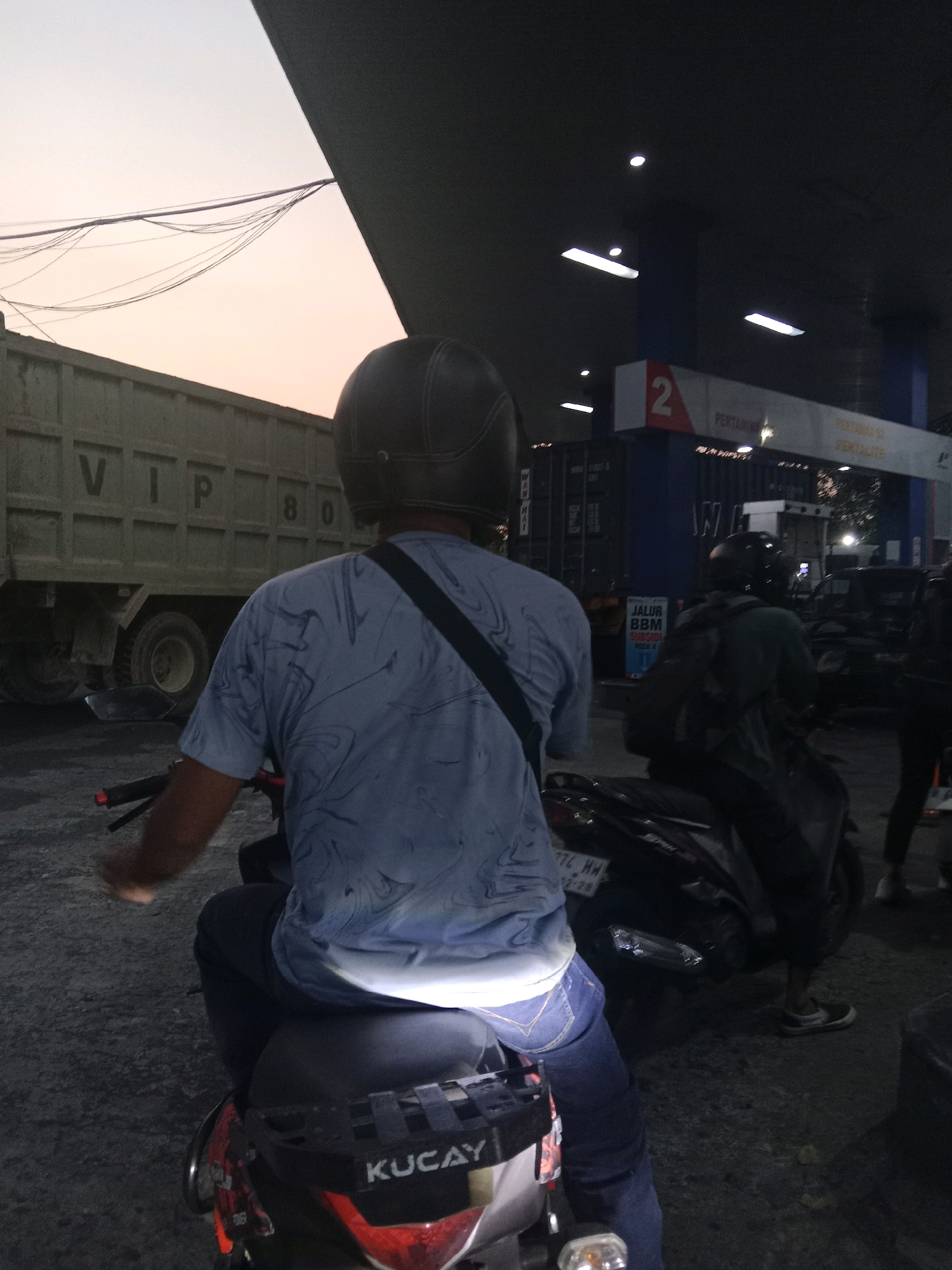 A person on a motorcycle, wearing a brown helmet and a light blue shirt with a dark swirl pattern, is parked at a gas station. They are facing away from the camera, looking towards the right. In the background, a large dump truck with the letters "VTP 80" is visible. The gas station has a blue and white sign with the number "2" and the word "PERTAMINA" on it. The sky is a light pink, suggesting it is either sunrise or sunset. Another person on a motorcycle, wearing a dark helmet and a dark jacket, is visible to the right of the main subject. The ground is asphalt, and there are some streetlights and wires visible. The image appears to be taken in Semarang, Indonesia, indicated by the presence of a Pertamina gas station, which is a common Indonesian state-owned oil and gas company.