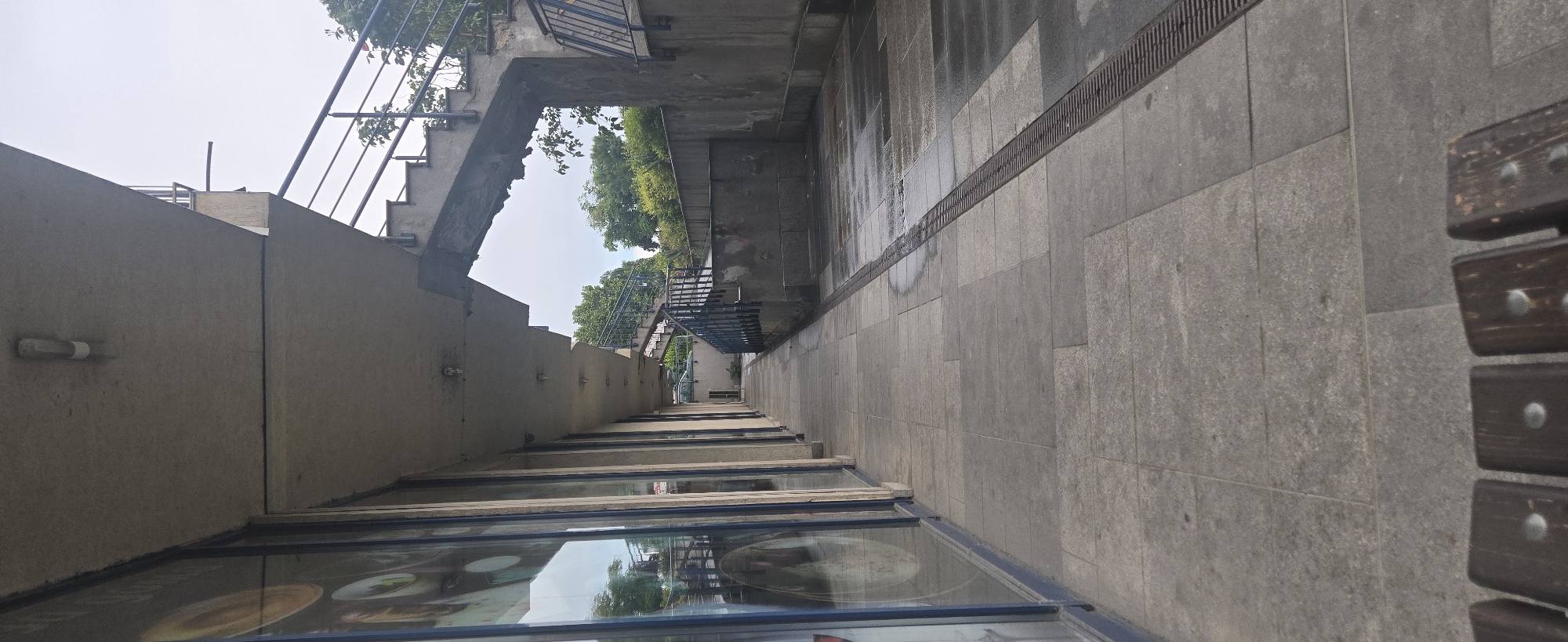 This image captures an empty urban pedestrian walkway or passage, possibly an underpass or a path alongside a building complex. The perspective is looking down the length of the path, which is paved with rectangular grey tiles.
On the left side of the path, a tall, textured concrete wall runs parallel to the walkway. Sections of concrete stairs with metal railings are integrated into or ascend alongside this wall, leading to an upper level where trees and the bright, overcast sky are visible. Small, horizontal light fixtures are mounted on the concrete wall. Along the base of this wall are large display windows, possibly shopfronts or advertising panels, reflecting the sky and distant greenery, though no legible text or specific products can be identified.
On the right side, the tiled ground shows distinct wet patches, suggesting recent rain or moisture. A long, dark metal drainage grate is embedded in the paving, running along the base of a darker, possibly wet, vertical surface. Further down the path on this side, another set of concrete stairs with metal railings can be seen. In the immediate foreground on the right, a portion of a dark wooden bench with prominent metal studs is visible.
No people or specific activities are depicted in the scene, which appears quiet and still. The time of day is daytime, indicated by the overall brightness, with an overcast sky suggested by the diffuse light and lack of harsh shadows. The architectural elements—concrete structures, metal railings, and tiled paving—point to a modern, urban setting.