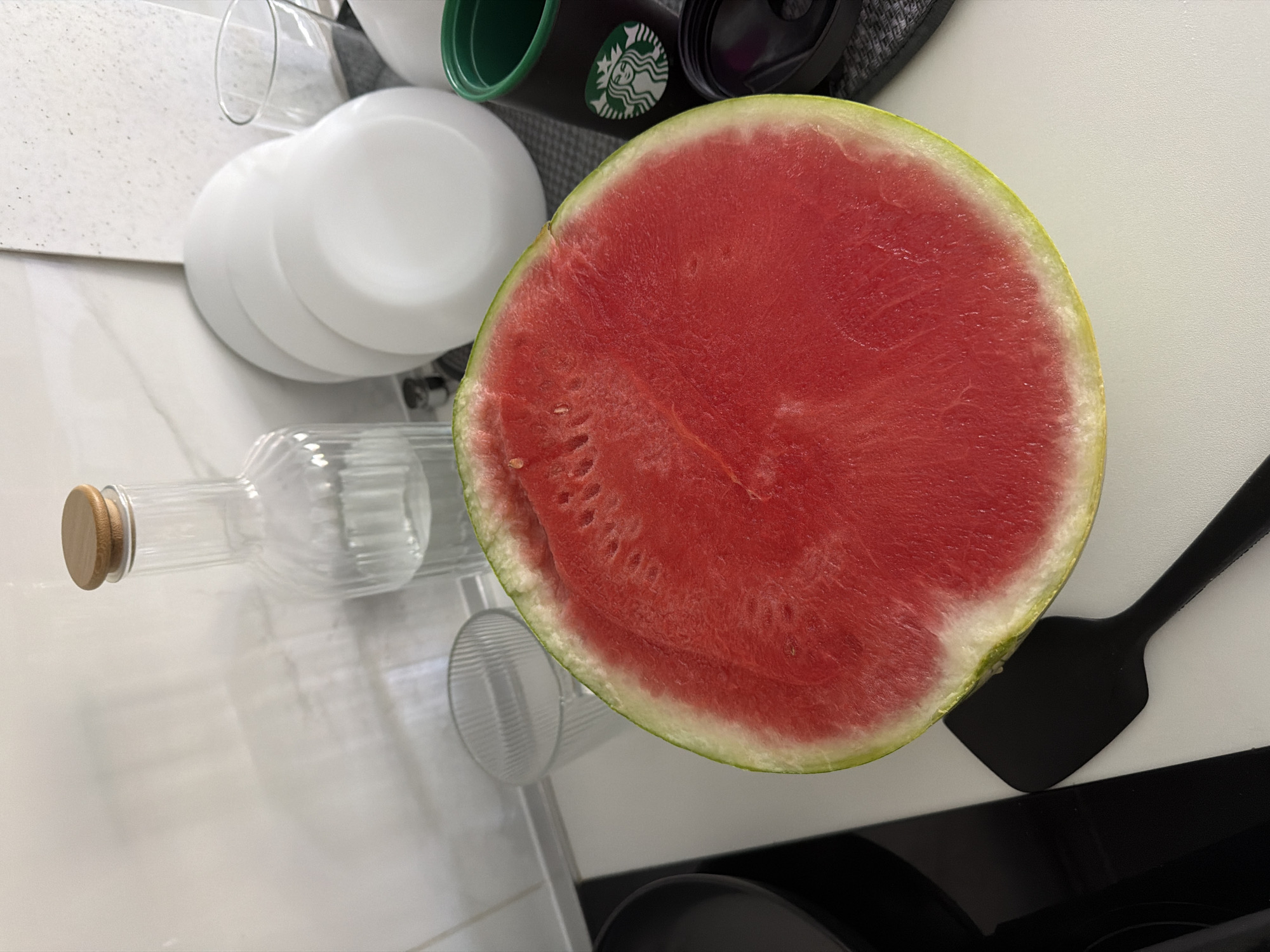 The media file depicts a close-up view of a half-cut watermelon on a white countertop. The watermelon is vibrant red with visible seeds and a pale green rind. To the left of the watermelon, a clear glass bottle with a wooden cork is positioned. Behind the bottle, a stack of white plates is visible. Further back, a dark Starbucks tumbler with the iconic siren logo is present. A black spatula is also visible to the right of the watermelon. The scene appears to be indoors, likely in a kitchen or dining area, given the presence of kitchenware. The lighting suggests it might be daytime, possibly in the morning or afternoon. There are no people visible in the image. The overall impression is that of food preparation or serving. The location context provided is Finestrat, Spain, though no specific visual cues in the image directly confirm this location.