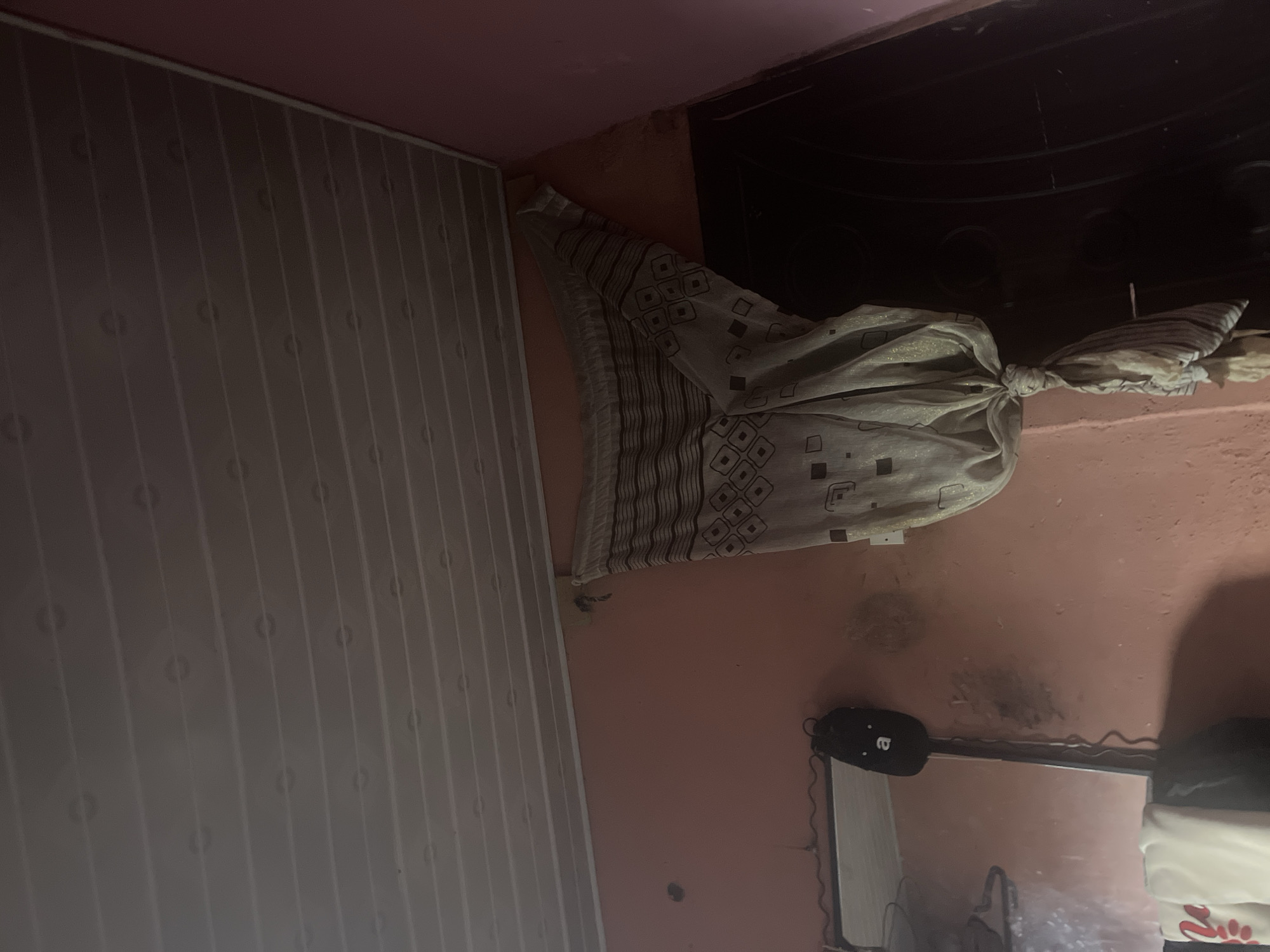This is a photograph taken indoors, likely in a residential setting in Jos, Nigeria. The room appears to be dimly lit, suggesting it could be nighttime or a poorly lit interior. The dominant colors are pink on the walls and a muted pattern on a fabric item hanging from the ceiling.
The most prominent object is a piece of patterned fabric, possibly a curtain or a sheet, tied in a knot and hanging from what seems to be the ceiling or a high fixture. The fabric has a geometric pattern of squares, some of which contain smaller squares. There are also striped sections on the fabric. To the left of the fabric, a section of a white, ribbed wall or ceiling panel is visible. The wall on the right is a plain pink, with some dark smudges or stains, indicating wear or dirt.
Further down on the right side of the image, a black object with a wire is attached to a reflective surface, which appears to be a mirror. The object has a face-like design with two dots for eyes and a curved line for a mouth, possibly a decorative item or a small appliance. Below the mirror, a part of what looks like a white garment with red lettering is visible.
The scene is static, with no discernible activity taking place. The overall impression is one of a cluttered or simply decorated living space, possibly a bedroom or a common area. The lighting and the state of the walls suggest it's a functional rather than a pristine environment. No people are visible in the image. No text is legible except for the possible red lettering on the white garment.