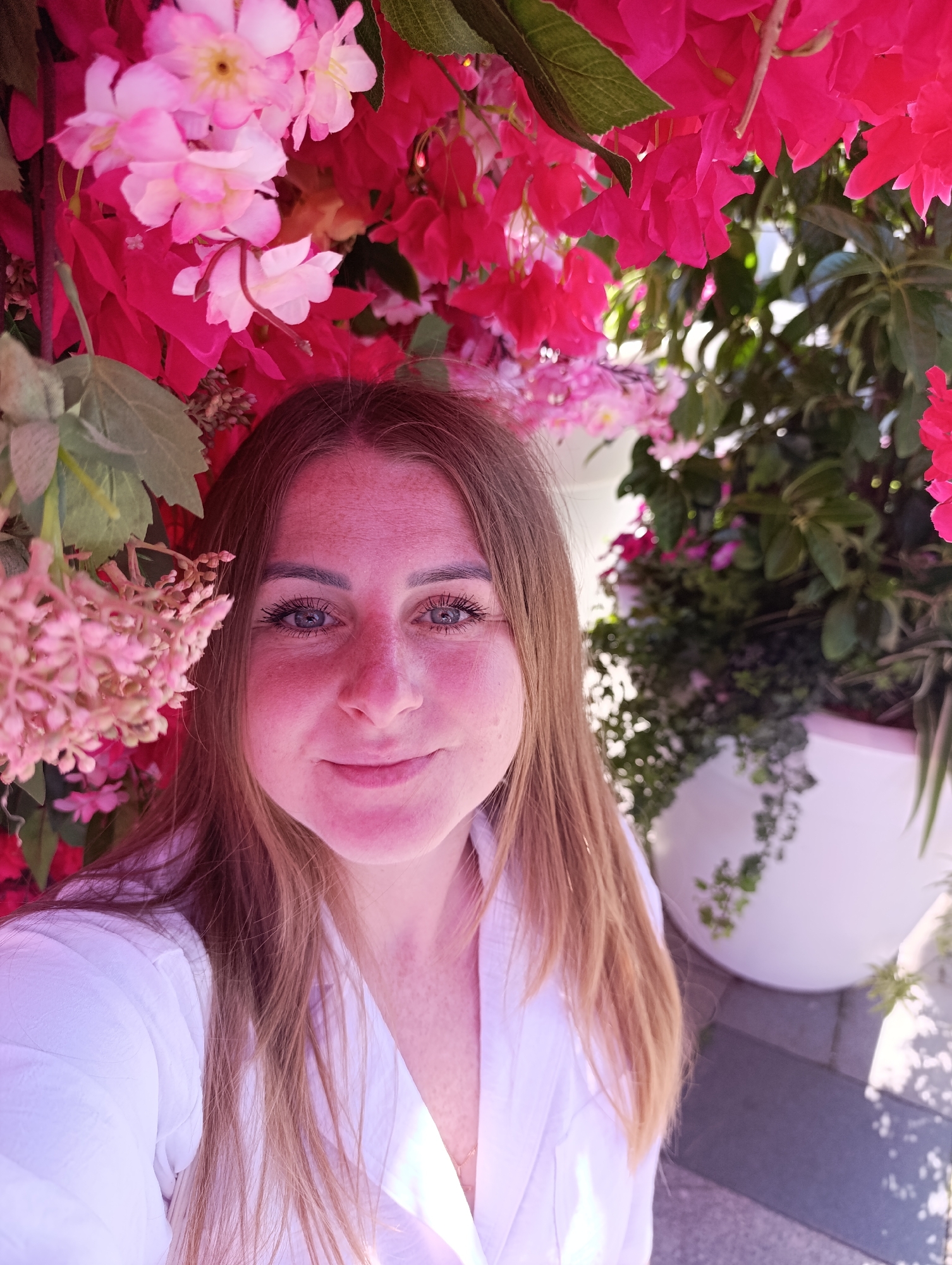 A female subject with long, light brown hair, blue eyes, and visible freckles on her nose and cheeks occupies the central foreground, gazing directly at the viewer with a slight smile. She is wearing a white, collared shirt with a v-neck, and a delicate gold chain is visible around her neck. Her head and upper torso are visible, cropped at the chest.
She is situated outdoors amidst a dense display of vibrant pink and fuchsia flowers, some resembling bougainvillea, which form a canopy above and around her. To the left, a cluster of smaller, lighter pink blossoms with visible buds hangs near her face. Interspersed among the flowers are green leaves.
In the background to the right, additional green foliage is visible, some contained within a large, rounded white planter. The scene is brightly illuminated by natural light, casting distinct shadows on a grey paved surface, possibly cobblestones or tiles, visible at the bottom right. The specific location is in the city of Unknown, Unknown.