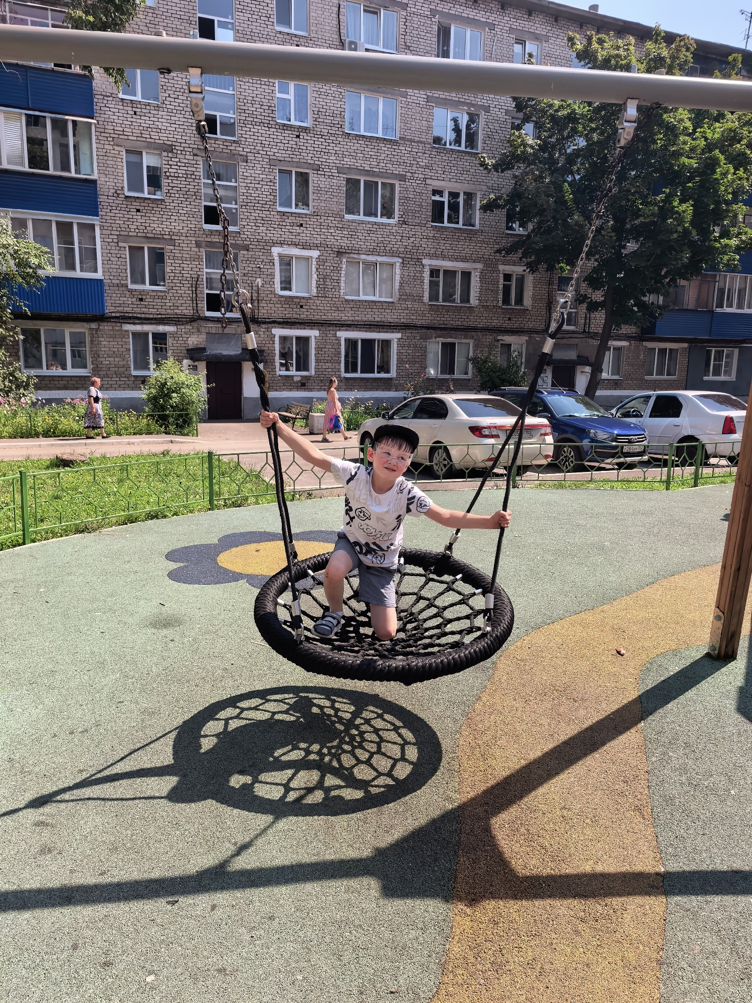 In a city playground, a young boy with short brown hair and glasses is seated on a black, circular rope swing. He wears a white t-shirt with a graphic print and grey shorts. The swing is suspended from a metal frame by two chains. The playground surface is a mix of green and yellow speckled rubber. In the background, a multi-story brick apartment building with numerous windows and blue balconies is visible. Several cars, including a white sedan, a blue sedan, and another white car, are parked on the street in front of the building. A green fence runs along the edge of the playground, separating it from a sidewalk and grassy area with flowers. Two women are walking on the sidewalk in the distance. The scene is illuminated by bright sunlight, casting shadows of the swing and its support structure on the playground. The image appears to be taken during the daytime in Sterlitamak, Russia, given the architectural style of the building and the parked vehicles.