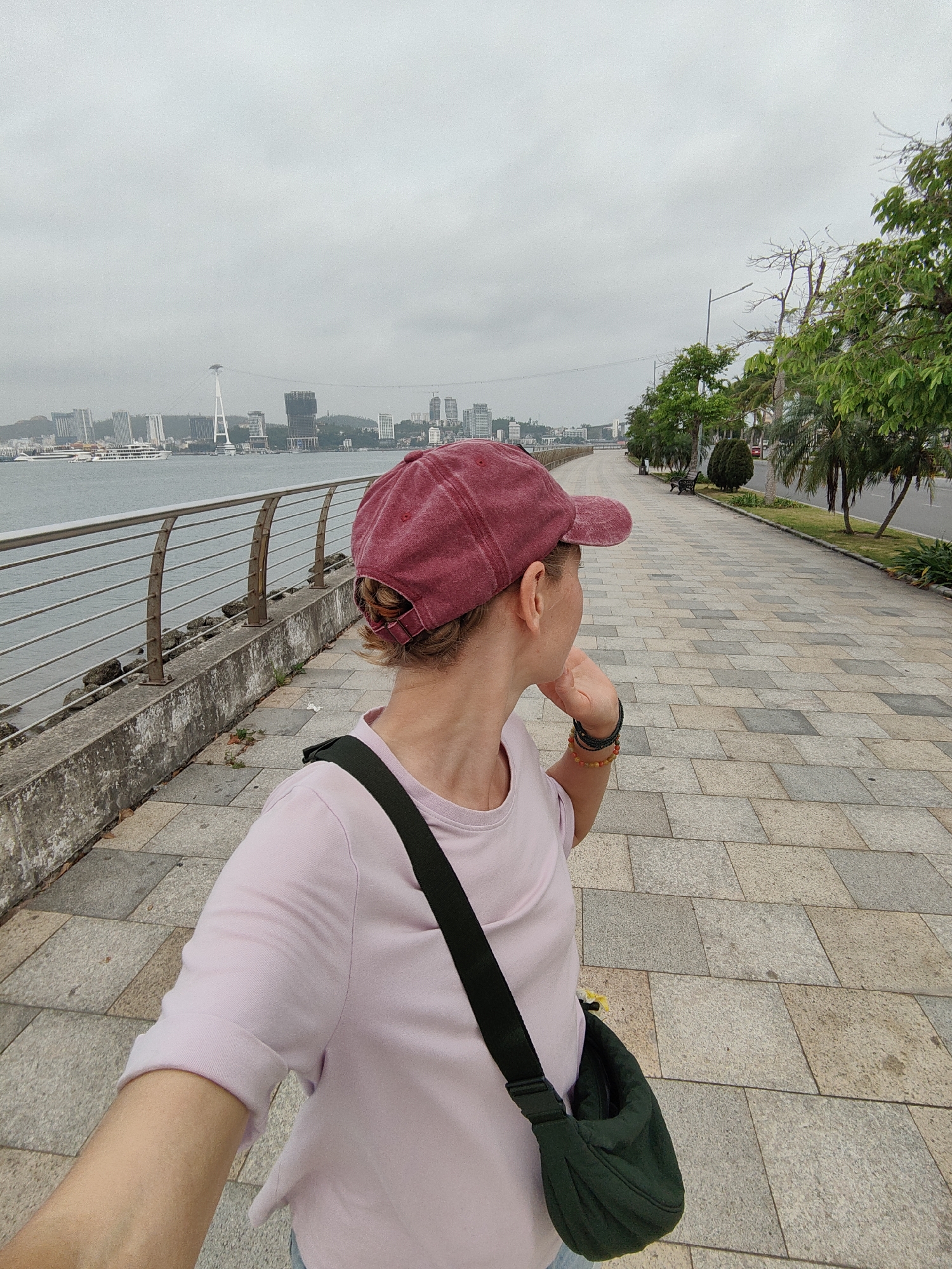 A first-person shot shows a person standing on a paved walkway next to a railing, facing a body of water and a city skyline. The person wears a pink t-shirt and a maroon cap, with a dark green bag slung over their shoulder. They are looking to the side, hand near their face. The walkway is composed of gray, rectangular paving stones. To the right of the walkway is a road with trees and benches. Across the water, a cityscape is visible under an overcast sky, including a cable-stayed bridge. The location is Hong Gai Ward, Vietnam.