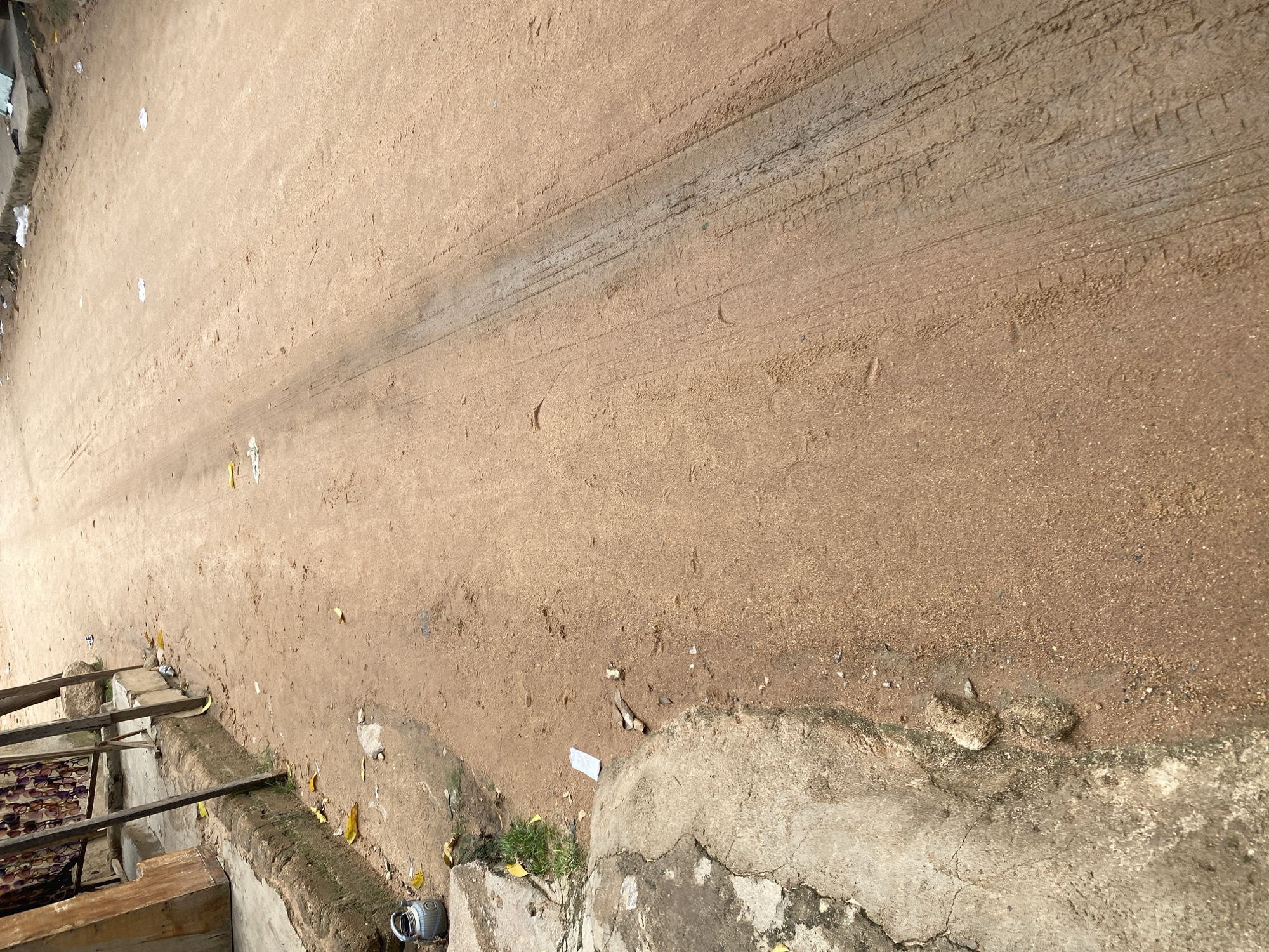 This image captures a close-up view of a dusty, unpaved surface, likely a dirt road or path, in Jalingo, Nigeria. The surface is a light brown, sandy material, showing distinct tire tracks from a vehicle. Scattered across the ground are a few small, light-colored pieces of debris, possibly paper or plastic, and some dry yellow leaves.
Along the left edge of the image, there is a raised concrete structure, possibly a low wall or embankment. Resting on this structure are wooden planks, forming a makeshift shelf or support. Below this, a rack holds multiple pairs of eyeglasses, suggesting a vendor or a display of goods. A small wooden crate or box is also visible.
To the right of the dirt surface, another concrete edge is present, appearing weathered and cracked in places. A small, metallic object, possibly a mug or pot with a handle, sits on this concrete edge near the bottom right corner.
The lighting suggests it is daytime, with shadows indicating the sun is at an angle. The overall impression is one of an outdoor, informal setting in a possibly rural or semi-urban area. There is no visible text in the image. The scene is static and lacks any explicit signs of human activity or strong emotional cues, focusing instead on the texture and elements of the environment.