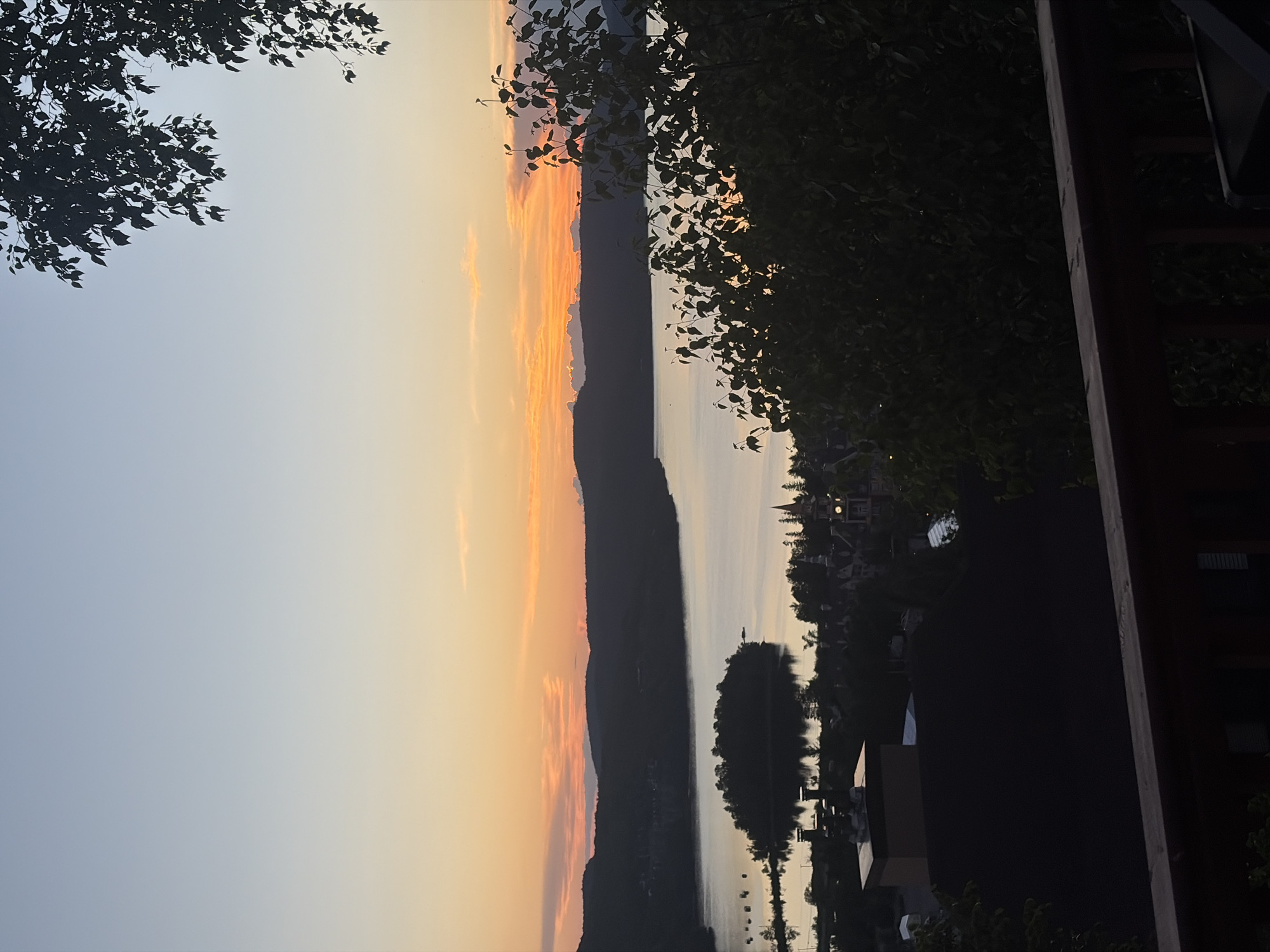 This is a photograph of a sunset over the city of Mont-Tremblant, Canada. In the foreground, a wooden railing and dark foliage obscure the lower portion of the image. The middle ground features a village with several buildings, including one with a distinctive turret and clock tower, and a large, dark-roofed building. A body of water, likely Lake Tremblant, is visible beyond the village, with a small, densely wooded island in the center. The background is dominated by silhouetted hills and a sky painted with shades of orange, yellow, and blue, indicative of sunset. The overall impression is of a serene, picturesque landscape at dusk.