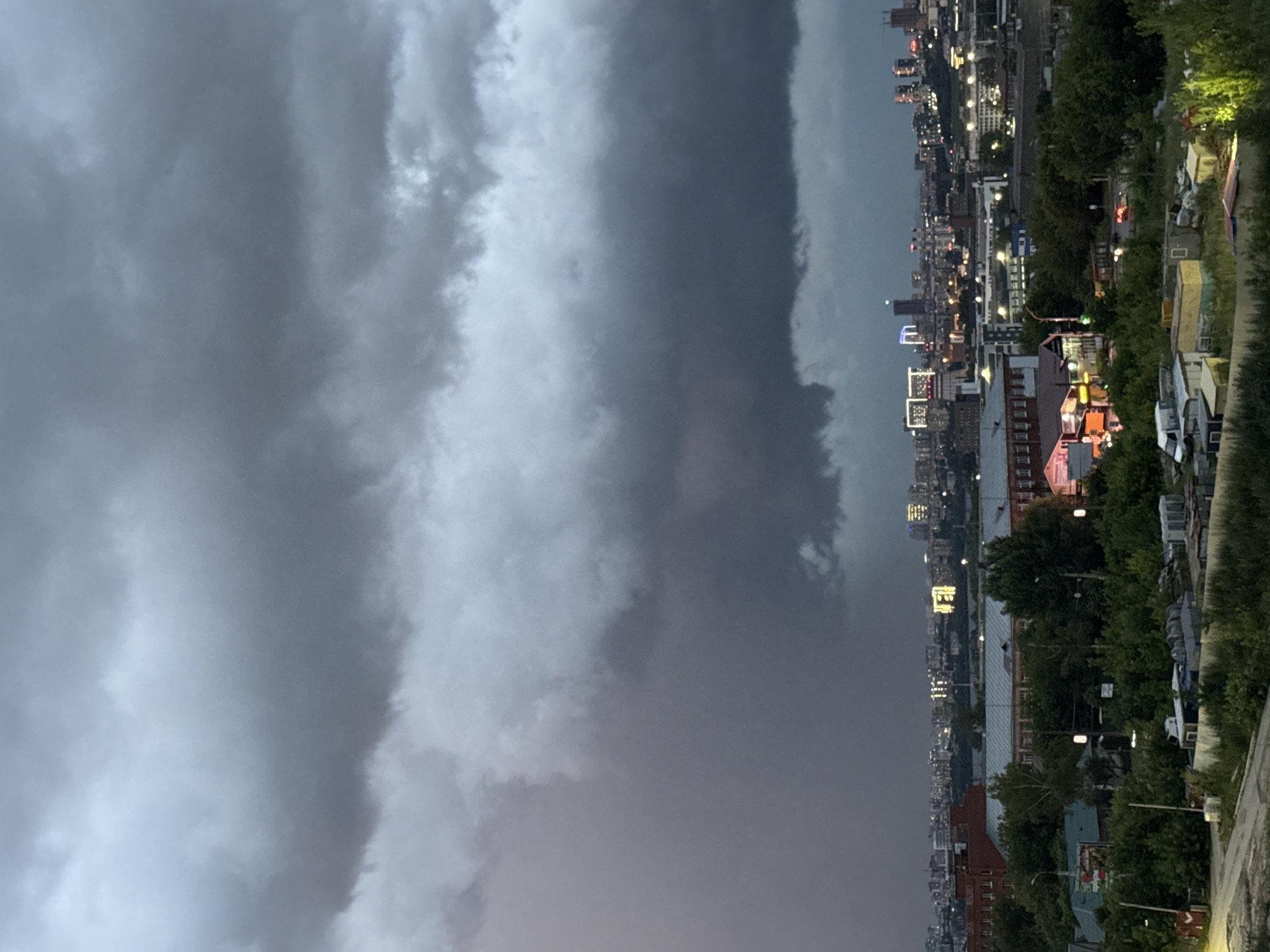 The image depicts a city skyline at dusk, dominated by heavy, dark storm clouds. The city itself is a mix of industrial buildings, trees, and taller structures with illuminated windows, suggesting it is a populated urban area. In the foreground, there are what appear to be storage sheds and piles of materials. The overall mood is somber and overcast, indicative of an approaching storm. Based on the provided information, this scene is located in Izhevsk, Russia.