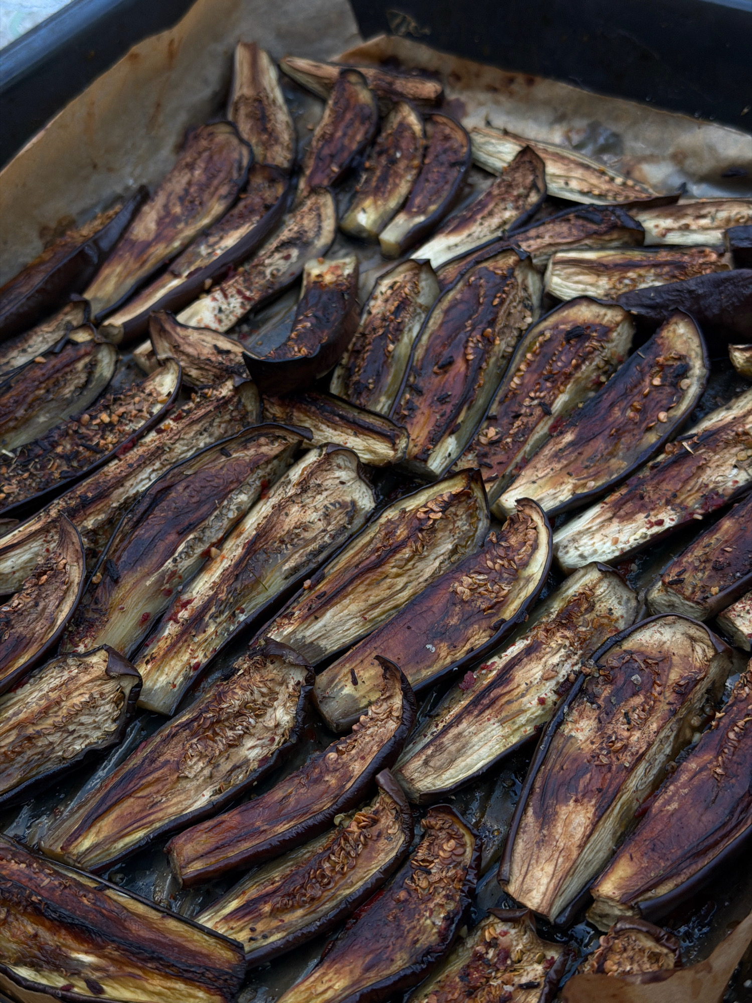The image displays a close-up, overhead view of numerous sliced eggplants arranged on a baking sheet lined with parchment paper. The eggplants are uniformly cut lengthwise and appear to have been roasted, exhibiting a deep brown, slightly charred coloration with visible softened flesh and seeds. Some pieces are seasoned with what appears to be red pepper flakes. The background is a dark baking tray. The scene is indoors and the lighting suggests artificial light or diffused natural light. The image does not contain any elements that would suggest a specific location in Novosilka, Ukraine.