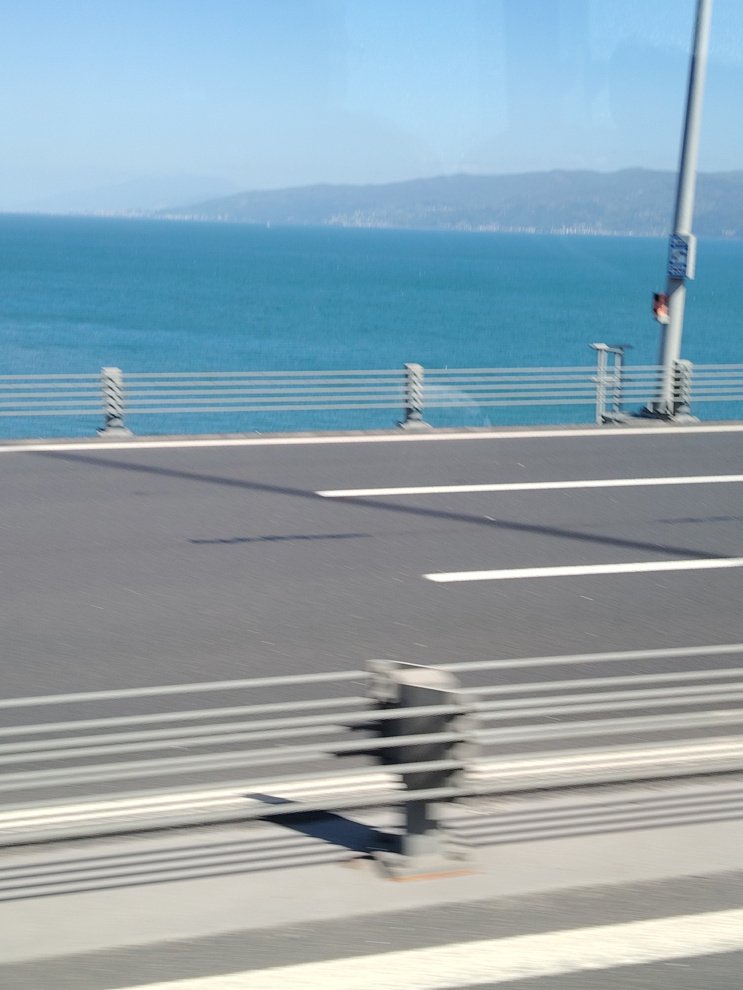 The image captures a bright, sunny view from a moving vehicle, likely crossing a bridge over a large body of water. The scene is dominated by a clear blue sky transitioning to a hazy horizon line, with distant mountains or land visible across the water.
In the foreground, a dark asphalt road with distinct white lane markings extends horizontally. The motion blur on the road surface and some foreground elements indicates the photograph was taken while in motion. To the right of the road, a metal barrier or guardrail with multiple horizontal bars separates the road from the open water. Several concrete posts with textured patterns support this barrier. A tall, light-colored utility pole stands on the right side of the road, near the barrier. On this pole, a small, blurry rectangular sign with a blue border and some white markings is visible, though the text is unreadable. Shadows from the barrier and possibly the vehicle itself are cast across the road surface.
The mid-ground features a vast expanse of calm, deep blue water, possibly a sea or a large gulf, reflecting the bright sky. Far in the distance, a long stretch of land or mountains appears as a muted, hazy blue-grey silhouette, suggesting a significant distance. Tiny, indistinct vertical shapes along the distant shoreline might be buildings or structures.
The time of day appears to be morning or early afternoon, given the bright sunlight and clear skies. The weather is fair and clear. The motion blur on the road and barrier, combined with the clear distant views, creates a sense of tranquil yet dynamic travel. Given the location context of Altınova, Turkey, this view is consistent with crossing a major bridge like the Osman Gazi Bridge over the Gulf of İzmit.