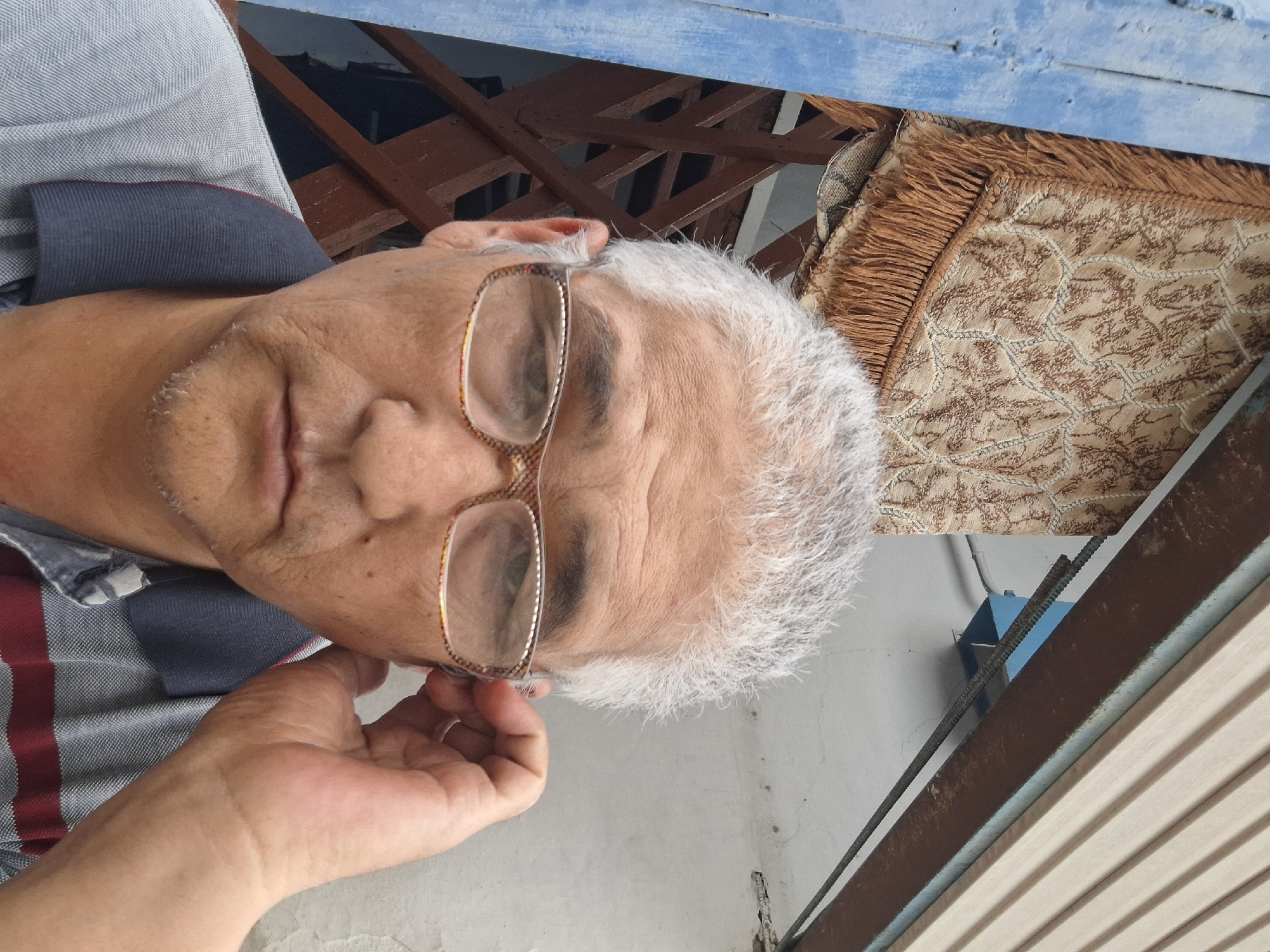 An elderly man with graying hair and glasses is taking a selfie outdoors. He is wearing a collared shirt with red and gray stripes. The background appears to be a weathered wall with some overhead structure. A patterned rug with fringe is visible hanging to the left of the man. There is a blue-painted wooden post also on the left. The scene suggests an outdoor residential setting, possibly a courtyard or a porch area, consistent with many older neighborhoods in Almaty, Kazakhstan. The man's hand is near his ear, suggesting he might be adjusting his glasses or listening. The lighting is natural, indicating it is daytime.