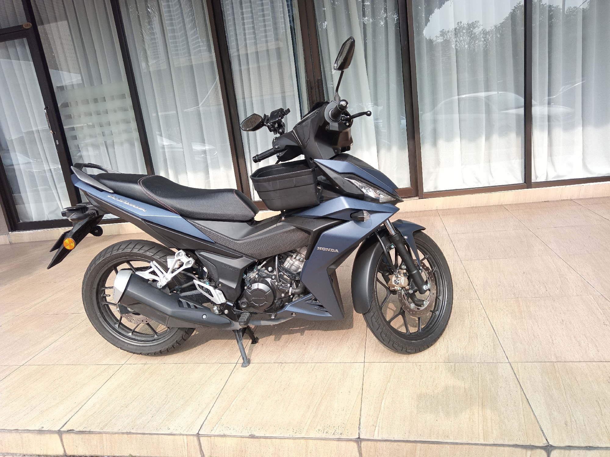 A dark grey Honda motorcycle is parked on a beige tiled patio in front of a building with large glass windows and white curtains. The motorcycle is a sport-style underbone model, featuring a front fender, handlebars with mirrors and a small black basket attached to the front, a black seat, and a silver exhaust pipe. The rear wheel and the lower engine components are visible. The building's architecture suggests it could be a modern commercial or residential property in Kuala Lumpur, Malaysia, given the style of the building and the reflective quality of the glass. There are no people present in the image. The environment is well-lit, indicating it is daytime.