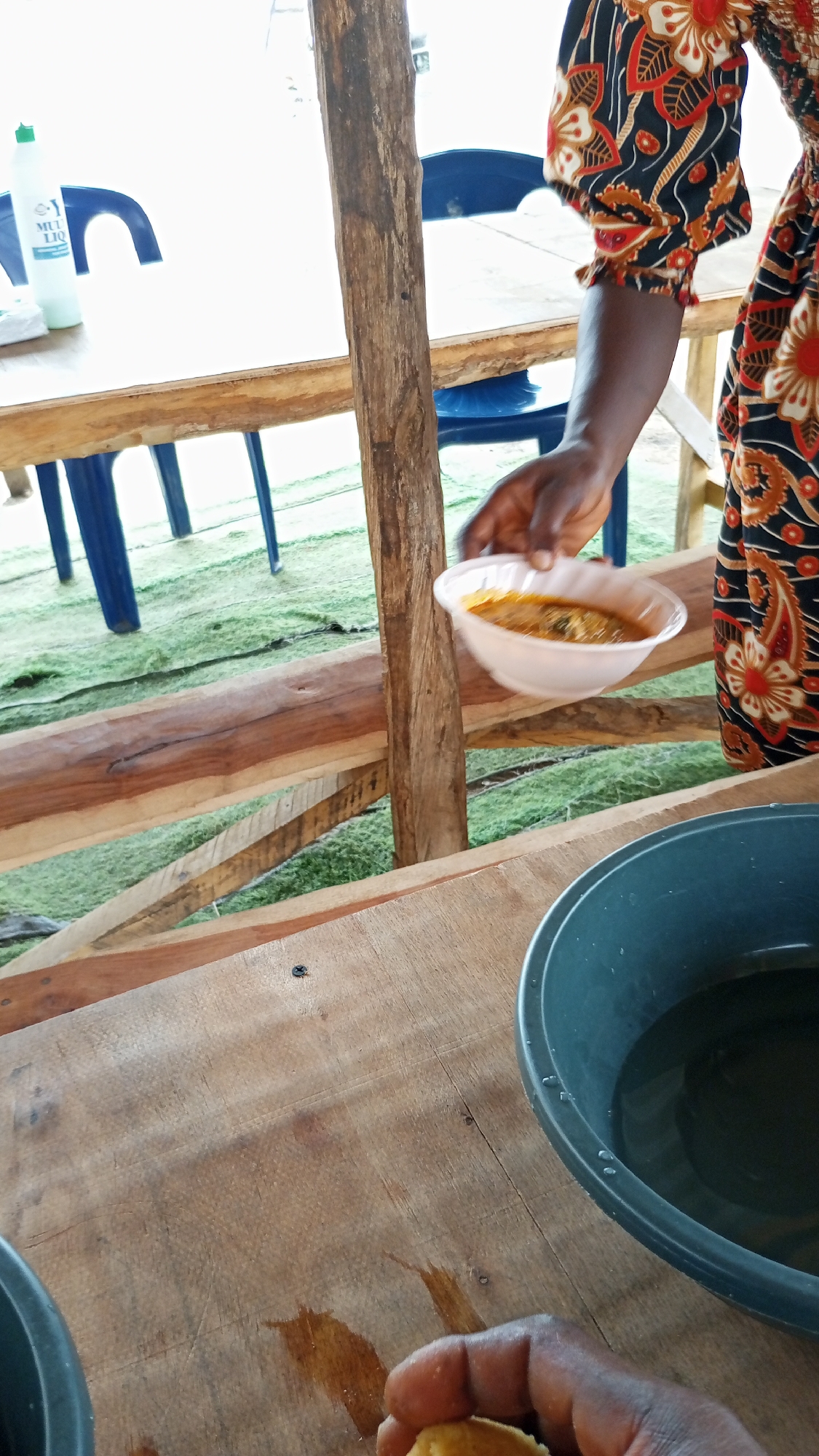 This vibrant image captures a moment in a rustic, open-air eating establishment, likely in Lokoja, Nigeria, as indicated by the context. The scene is bright, suggesting daytime.
In the foreground, a person's dark-skinned hand is visible on a light brown wooden table, holding a piece of light-colored solid food, commonly referred to as "swallow" in West African cuisine, which is typically eaten with stew or soup. Adjacent to this hand is a dark grey or black plastic basin filled with clear water, possibly for handwashing.
Above, another person's arm, also dark-skinned, extends from the right, holding a white disposable bowl containing an orange-brown stew or soup with visible green elements. This person is wearing clothing with a distinctive red, black, and orange floral and geometric pattern, reminiscent of West African Ankara fabric.
The eating area features simple wooden structures, including wooden tables and posts that support the roof (not fully visible). The ground beneath the tables is covered with a green textured mat or fabric. In the background on the left, a white plastic bottle with a green cap, partially obscured, is seen on a table. The visible text on the bottle reads "MUL LIQ," likely part of "MULTI LIQUID," suggesting it could be a cleaning agent or hand sanitizer. Blue plastic chairs are partially visible around the tables in the background.
The image depicts an everyday dining experience, highlighting the food and the functional, unpretentious setting.