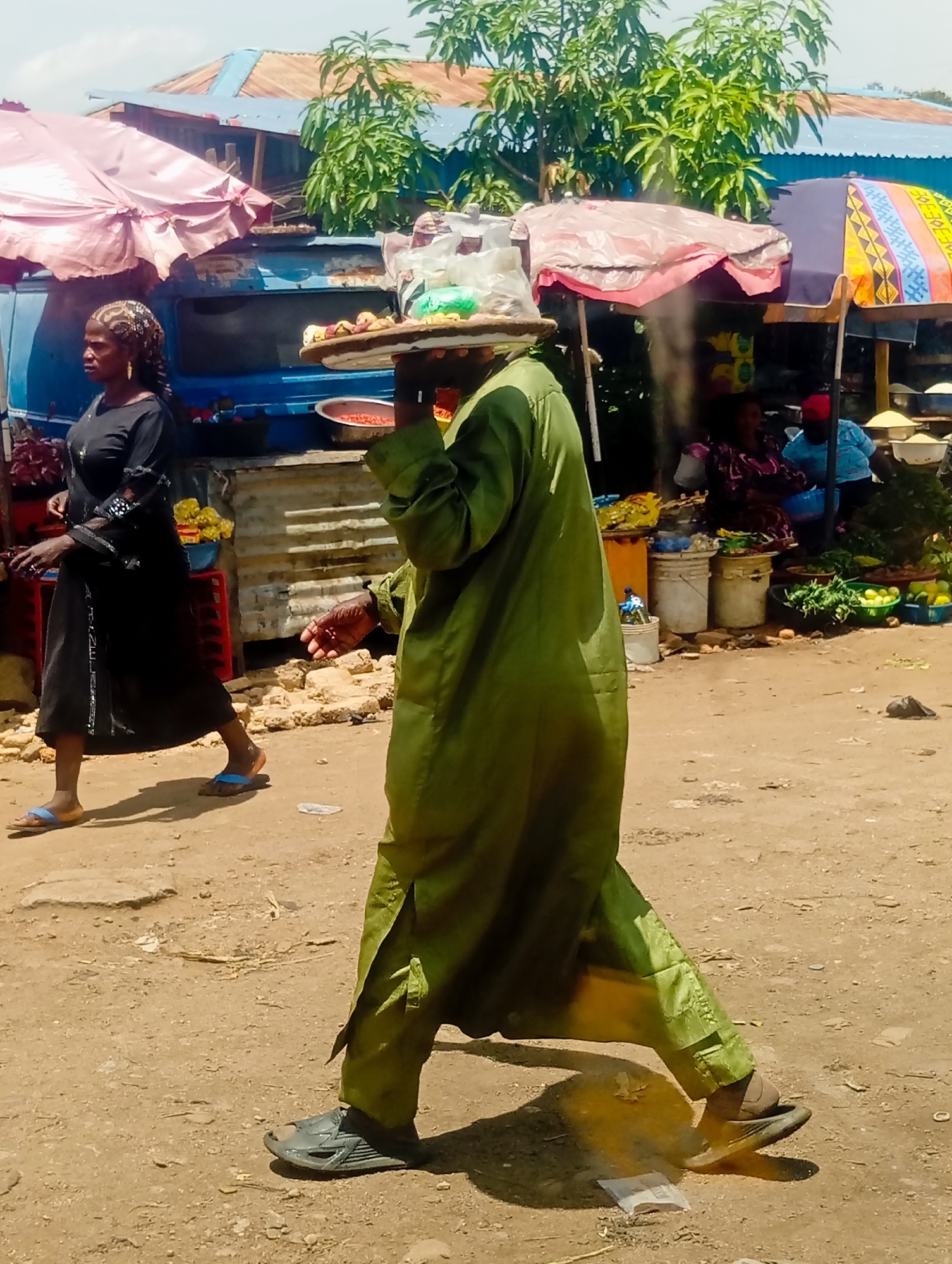 A busy outdoor market scene is depicted in Bukuru, Nigeria, under a clear sky. A man in a lime-green robe and pants walks with a tray of items balanced on his head. He is moving away from the camera, his right foot forward and casting a distinct shadow on the dusty ground. He wears dark, open-toed sandals. To his left, a woman in a black abaya and headscarf walks in the opposite direction, her left foot forward and her right foot slightly behind. She wears blue flip-flops and has a gold earring.
Behind them, a makeshift stall is set up with an assortment of goods displayed under colorful umbrellas. One umbrella is pink and draped over a blue structure, possibly a vehicle. Another umbrella is blue, purple, and yellow with a geometric pattern. Various vendors, mostly women, are visible behind the stalls, tending to their merchandise. Displays include bowls of red peppers, baskets of green vegetables like leafy greens and limes, and other unidentifiable food items. Buckets and crates are used to hold and organize the produce. The ground is unpaved and dusty, characteristic of a marketplace in a developing urban area. The overall impression is one of vibrant commerce and everyday life in a Nigerian market.