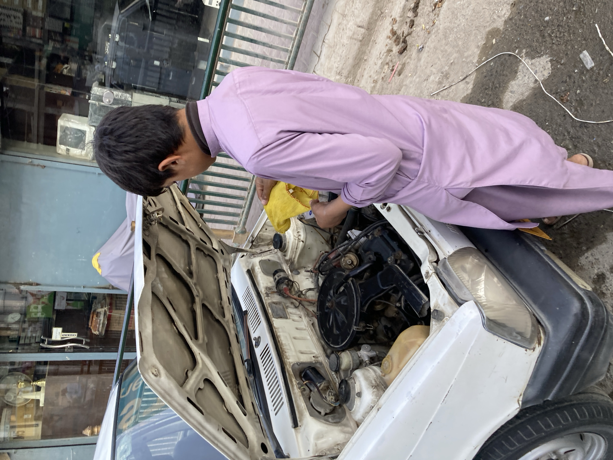 A person is bent over the open hood of a white car in what appears to be a city setting. The individual is wearing a long, light purple tunic-like garment and is holding a yellow cloth in their hands, seemingly working on the car's engine. The car has a black bumper and a visible headlight. The hood of the car is open, revealing the engine components. The backdrop features a building with glass windows showcasing various objects inside, and a metal railing is visible alongside a concrete pavement. The scene is in Quetta City Tehsil, Pakistan.