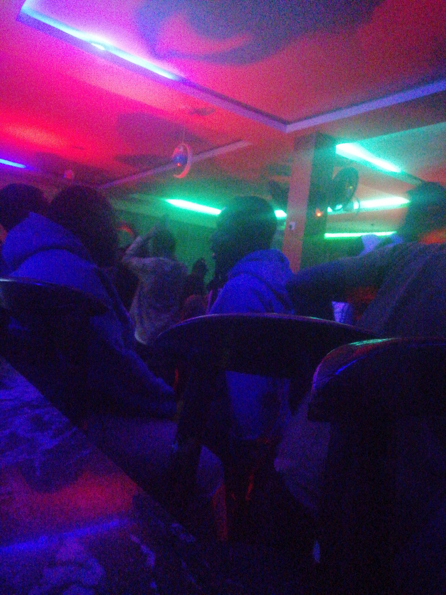 The media file displays an indoor scene, likely an entertainment venue such as a bar or club, located in Jos, Nigeria. The image captures a low-light environment illuminated by vibrant artificial lighting in shades of fuchsia, pink, blue, and bright green. Several people are visible from behind, their forms largely obscured by the darkness, suggesting they are seated in chairs or booths, facing away from the camera. They appear to be wearing dark clothing.
The ceiling features multiple linear fluorescent light fixtures, casting strong colored glows across the space. A small, reflective disco ball or similar circular ornament hangs from the ceiling in the mid-ground. The overall atmosphere is dark and vibrant, typical of a nighttime gathering or social event. The image quality is grainy, a characteristic often seen in low-light photography. No specific activity beyond gathering and likely socializing can be definitively identified. There is no visible text in the image.