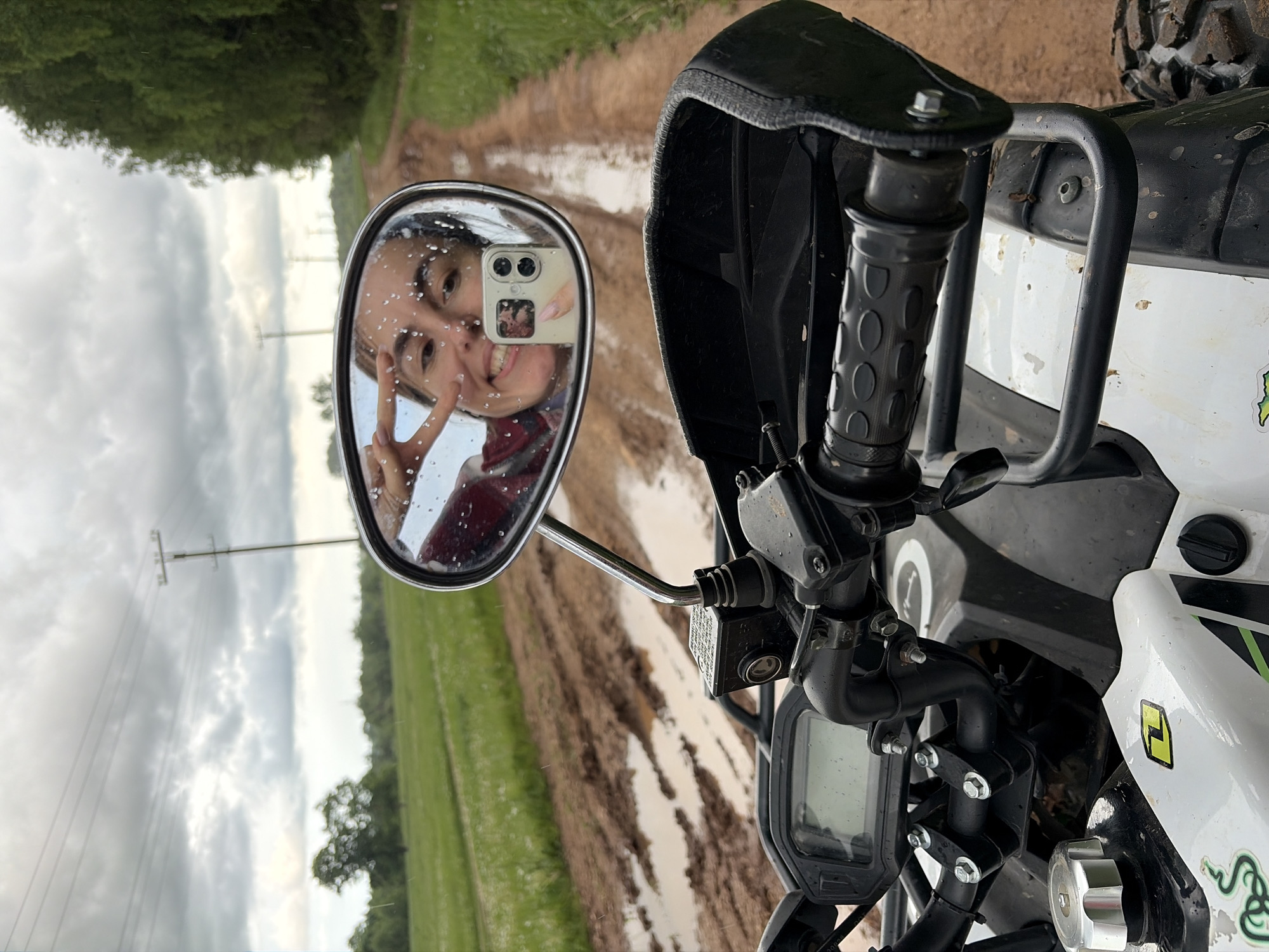 The image depicts a view from the front of an all-terrain vehicle (ATV). The focus is on the ATV's handlebars, gauges, and a side mirror. The mirror reflects a young woman taking a selfie, holding up a peace sign. Water droplets are visible on the mirror's surface. Behind the ATV, a muddy dirt road stretches into the distance, flanked by green fields and trees. Utility poles are also visible in the background. There is no indication of the location.