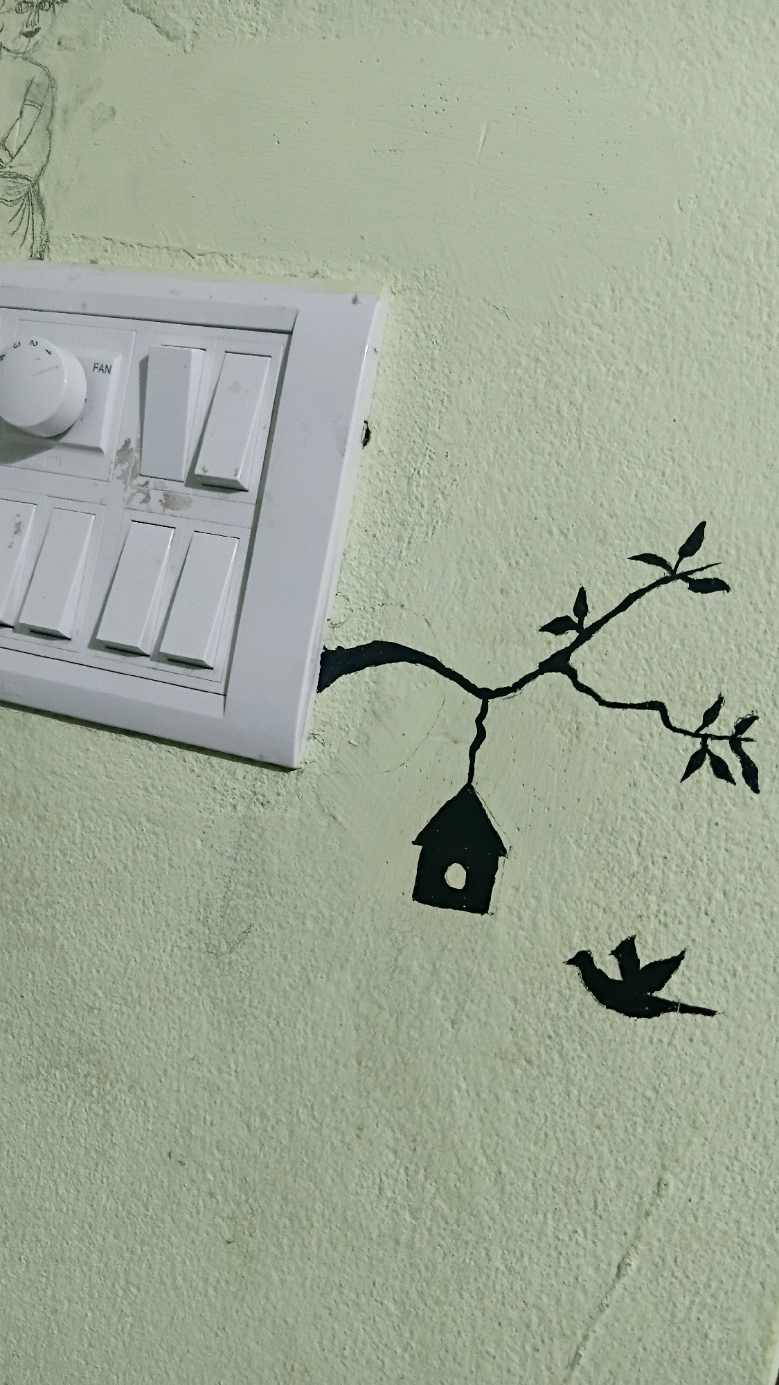 The image shows a light green wall with a white electrical switchboard on the left. The switchboard has multiple switches and a rotary dial labeled "FAN". To the right of the switchboard, a black silhouette painting on the wall depicts a tree branch with leaves, a birdhouse hanging from the branch, and a bird in flight. The environment appears to be indoors, likely a domestic setting. The drawing of a human figure is faintly visible on the upper left of the wall, partially obscured by the switchboard. This scene is located in Kistapur, India.