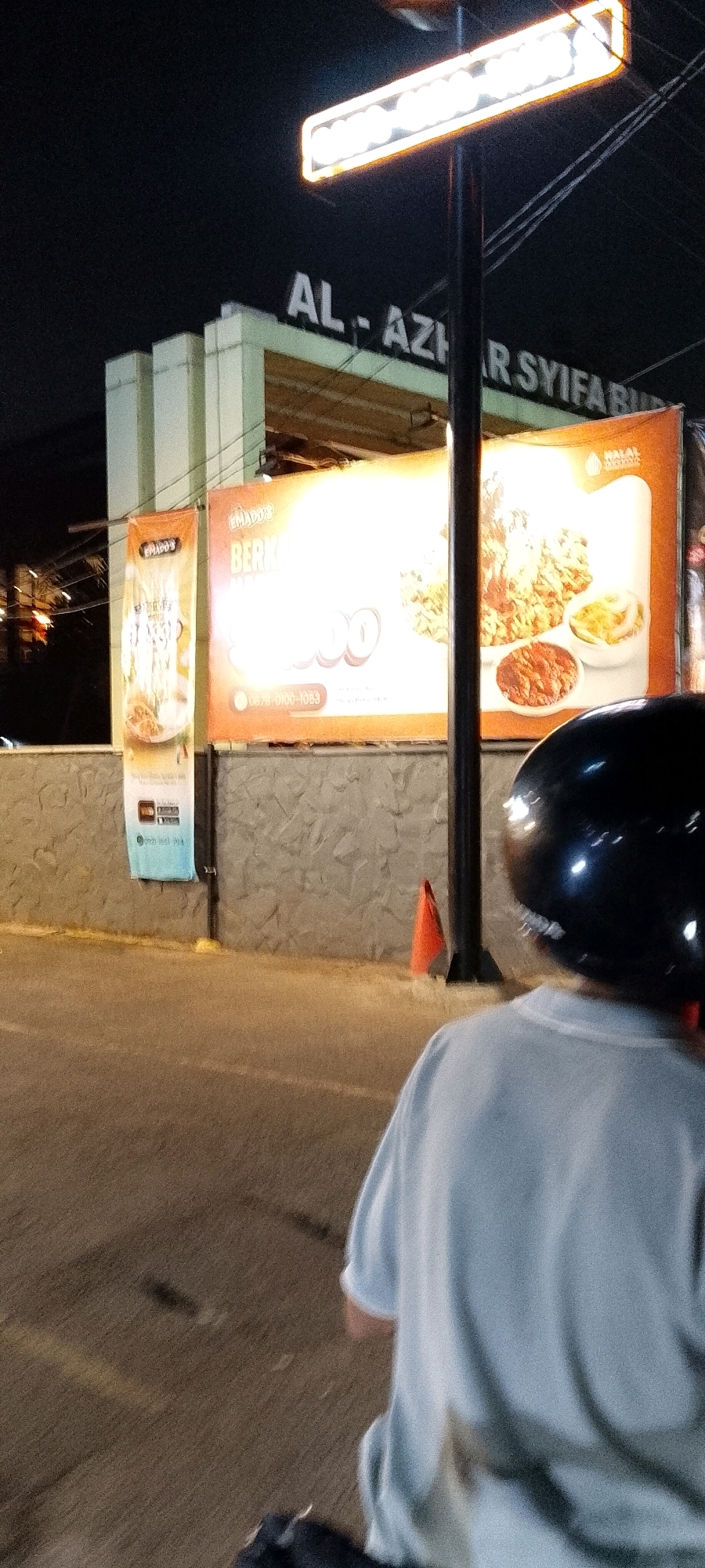 In Gunung Putri, Indonesia, at night, a person wearing a light blue t-shirt and a black helmet is on a motorcycle, facing away from the camera. The background shows a building with a sign reading "AL-AZHAR SYIFA BUDHI" in white letters. Adjacent to the building are two large banners. The right banner, illuminated, displays images of food, including fried chicken and chili sauce, with text that appears to be in Indonesian. The left banner, also vertical, shows images related to food as well, and some text. A lamppost with a bright rectangular light is positioned in front of the building. An orange traffic cone is visible on the right side of the image, near the motorcycle. The ground is paved, and the overall atmosphere suggests an urban setting at night.
