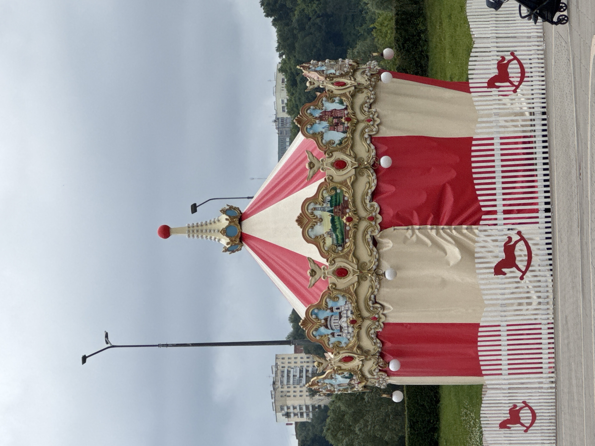 The image shows a carousel with a red and cream striped roof, decorated with ornate golden trim featuring small paintings of buildings. The carousel is surrounded by a white picket fence adorned with red rocking horse silhouettes. In the background, a wooded hillside with buildings can be seen, suggesting an urban or suburban setting. The sky is overcast and gray. To the left of the carousel, a tall lamppost is visible. The scene appears to be an outdoor public space in Nizhny Novgorod, Russia, possibly a park or a recreational area.