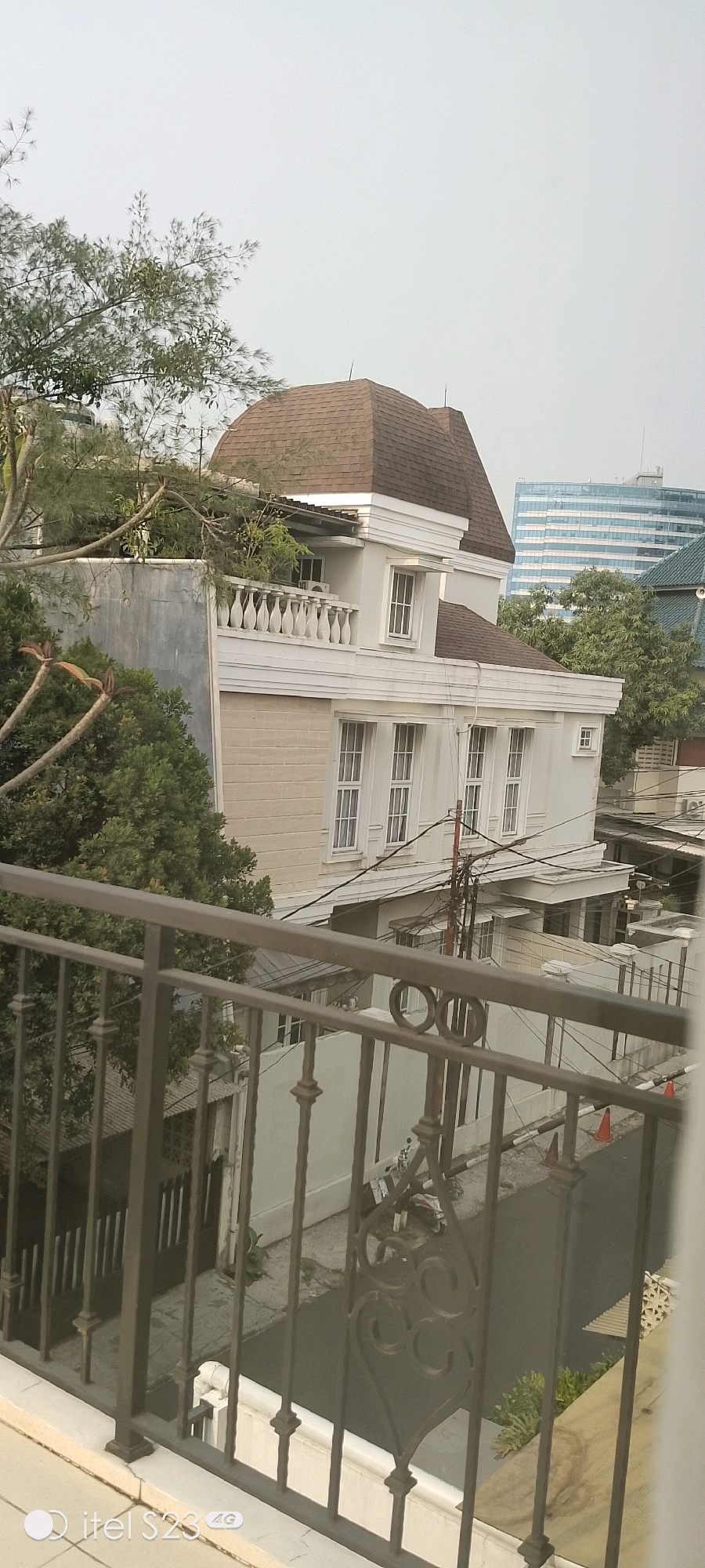 This is a medium-shot, eye-level, outdoor photograph captured on an overcast day, likely in South Jakarta, Indonesia. The foreground is dominated by a dark metal railing, possibly from a balcony, with decorative elements including circles and scrollwork. Beyond the railing, a street is visible, paved with asphalt and lined with a low concrete wall.
The primary subject of the image is a white, multi-story house with a distinctive brown, curved roof that resembles a dome or a rounded turret. The house features numerous white-framed windows on multiple levels, some of which appear to be casement windows with multiple panes. A white balustrade runs along the top of a lower section of the house, overlooking the street. Green trees with sparse foliage are visible around and behind the house, some branches extending over the roof. To the right and behind the house, a tall, modern building with a blue glass facade is visible, suggesting an urban environment. Electrical wires and utility poles are present in the mid-ground. There are no people clearly visible in the frame. The overall atmosphere is somewhat muted due to the cloudy sky.