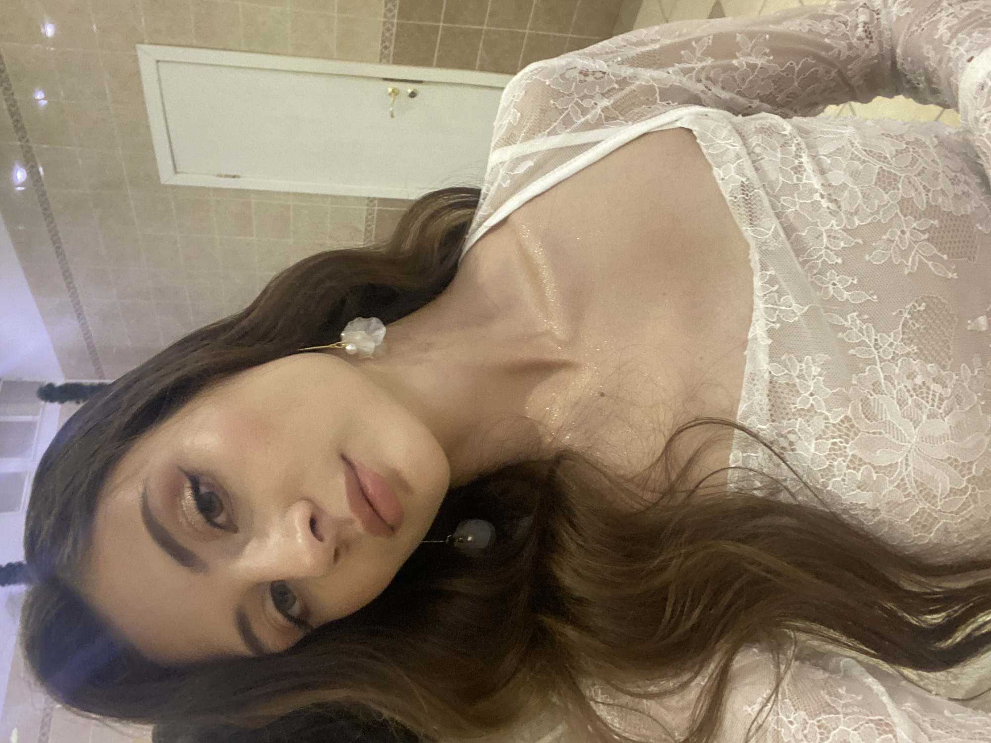 This media file is a close-up, sideways shot of a young woman lying down, with her head tilted. She has long, wavy, brown hair and is wearing a white, lacy top. Her makeup includes eye shadow and lipstick, and she is wearing delicate earrings. Her skin appears to have a shimmering highlight, possibly from glitter or a luminous lotion.
The background suggests an indoor setting, possibly a bathroom or a room with tiled walls and a white cabinet or door. The lighting is dim, creating a moody atmosphere. The overall impression is one of intimacy and perhaps a moment of quiet reflection or preparation.
There is no visible text in the image, and the time of day and weather are not discernible. The location context provided places this scene in Votkinsk, Russia.