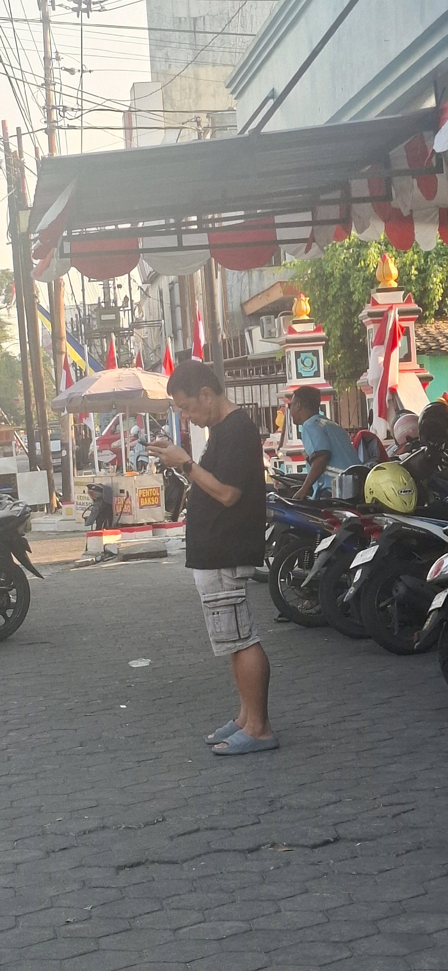 The image depicts a street scene in Surakarta, Indonesia, during daylight. A man of Asian descent, wearing a black t-shirt, light-colored cargo shorts, and blue slip-on sandals, stands facing a mobile device in his hands. He is positioned in the foreground, to the left of center. Behind him, to the right, another man in a blue t-shirt is partially visible, leaning over a motorcycle. The background features a street lined with parked motorcycles. A food stall with a red and white umbrella and signage that reads "PENTOL BAKSO" is also present. Decorative red and white banners are strung across the street overhead. Utility poles with numerous wires are visible on the left side of the image. The ground is paved with interlocking hexagonal bricks. The overall impression is that of a busy urban environment, possibly a market or roadside vendor area.